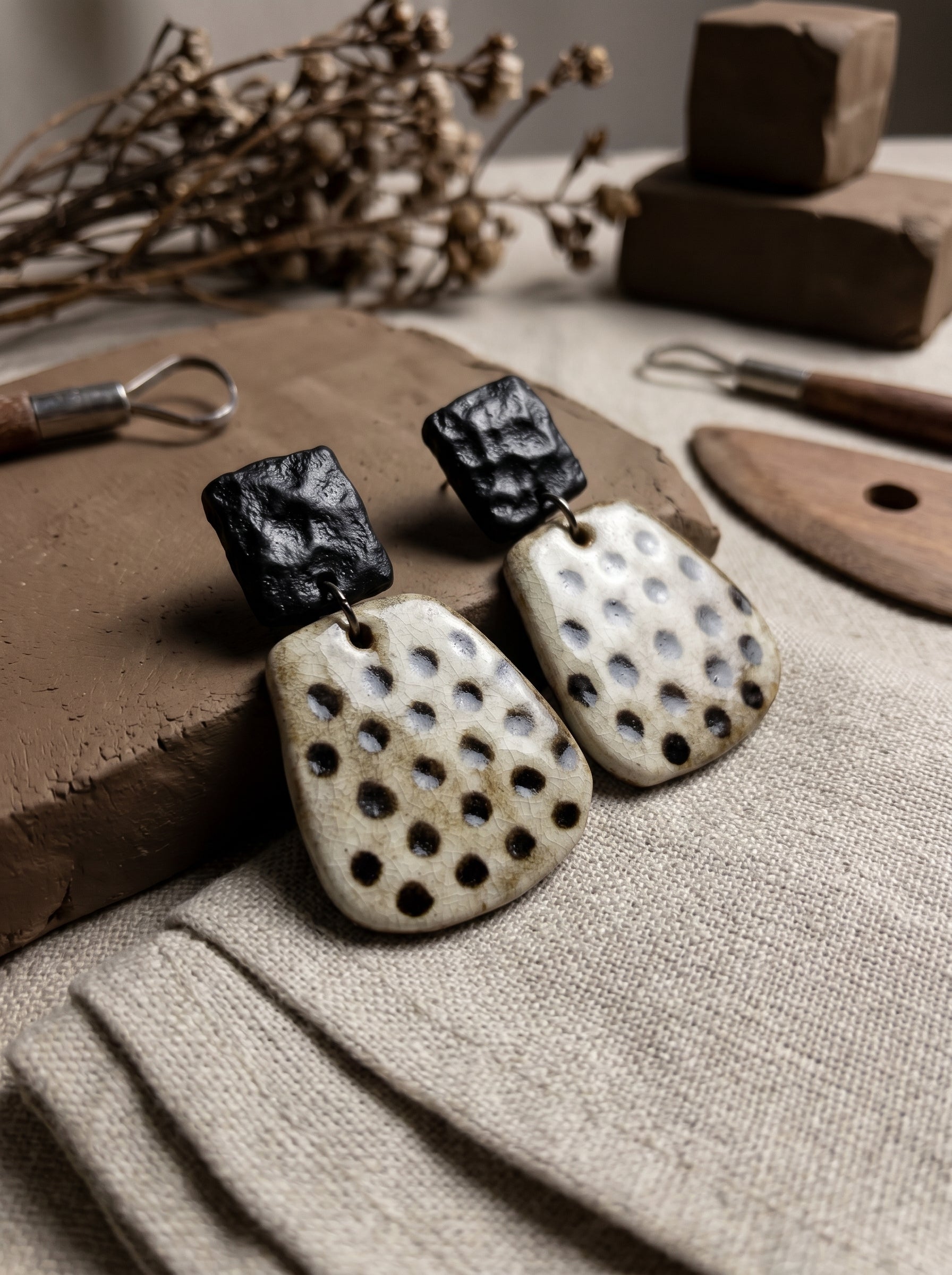 HECROWN speckled geometric ceramic earrings resting on raw clay with sculpting tools. Showcasing the authentic Jingdezhen handmade jewelry process and artisan focus.