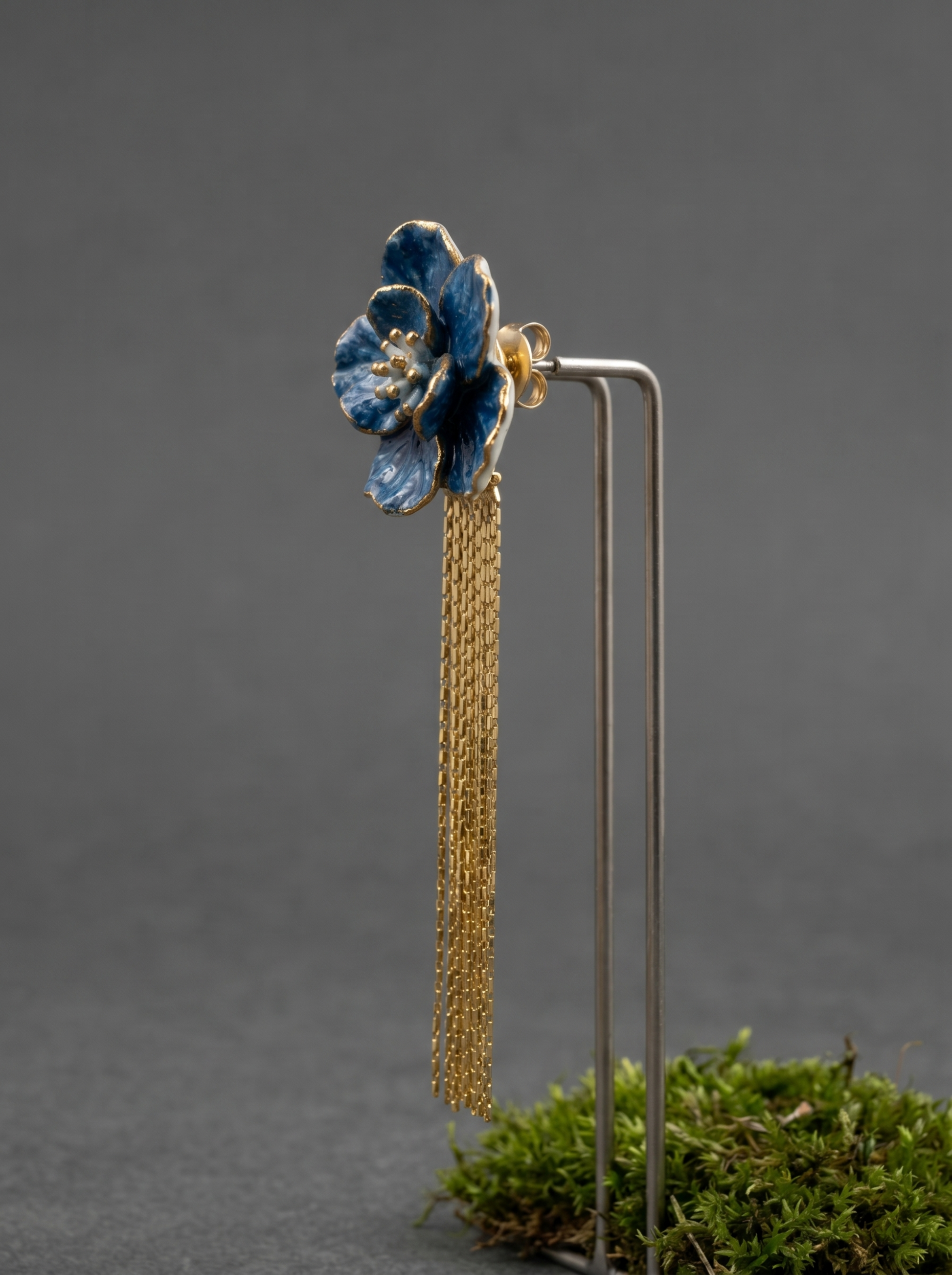 Side view of a blue porcelain flower earring with gold tassels on a minimalist stand with moss. Highlighting the sculptural Jingdezhen ceramic art and handmade jewelry quality.