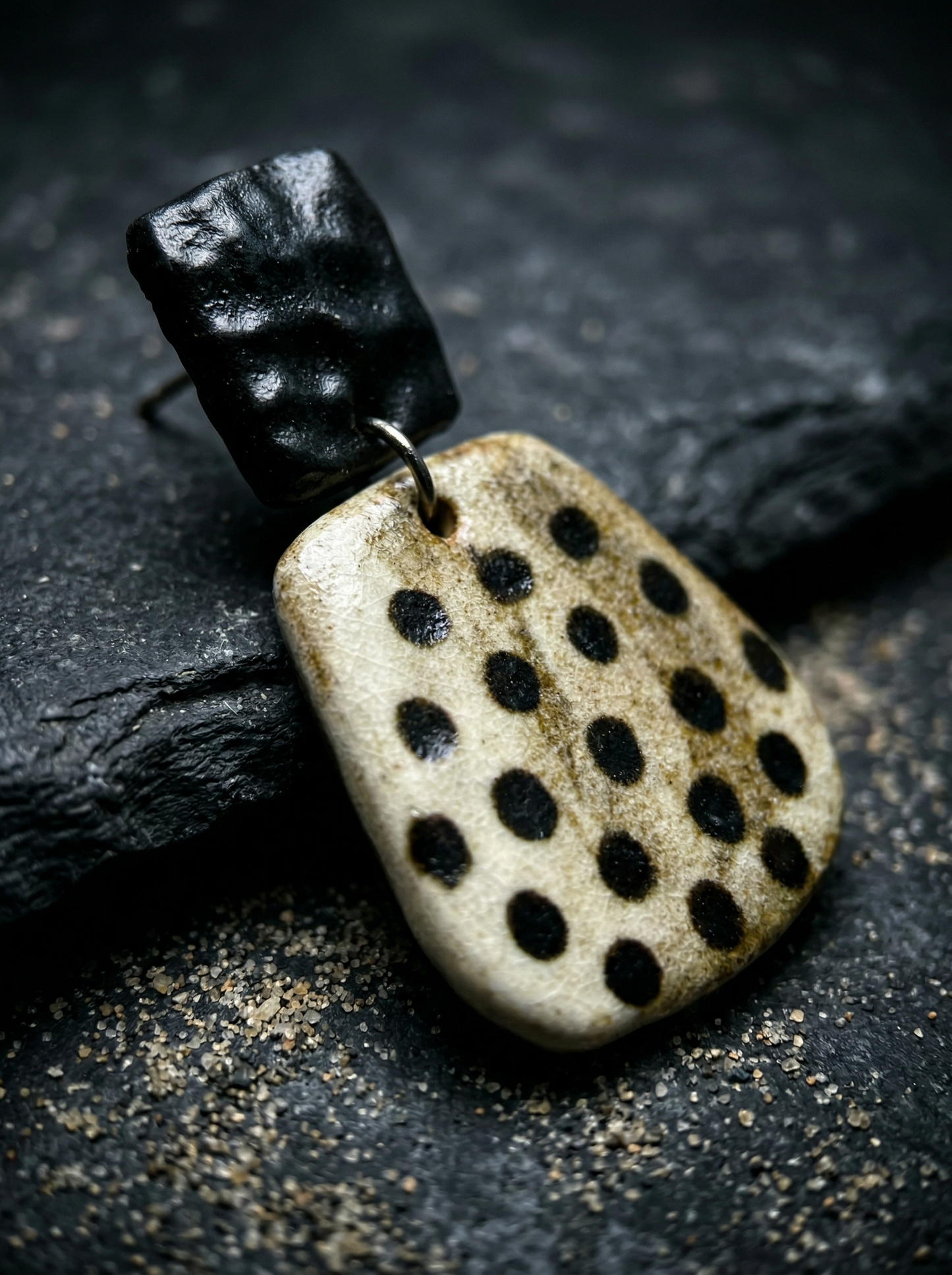 Close-up of a HECROWN speckled ceramic earring on dark slate with sand. Highlighting the hammered black top and rustic texture of true handcrafted artisan jewelry.