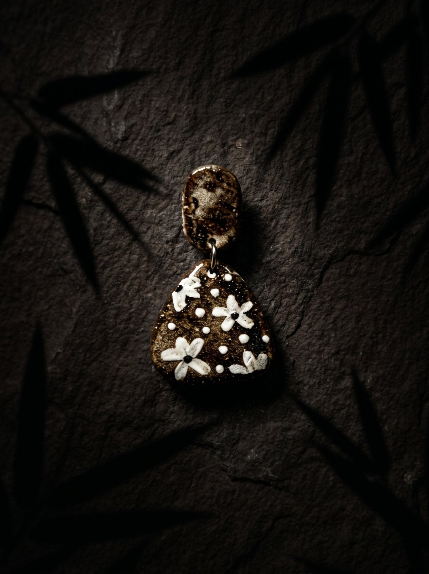 HECROWN dark ceramic dangle earrings featuring painted white botanical patterns, resting on dark slate under bamboo shadows. A unique artisan handmade jewelry gift.