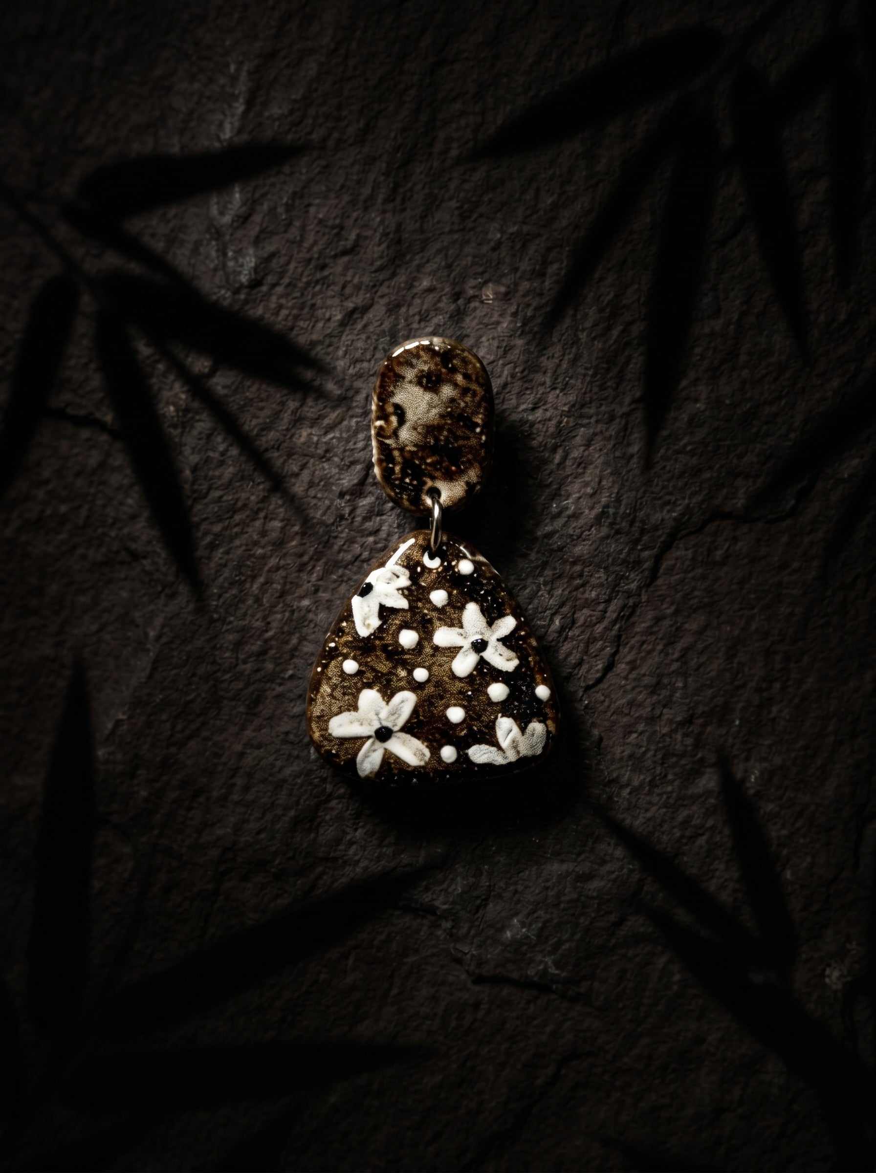 HECROWN dark ceramic dangle earrings featuring painted white botanical patterns, resting on dark slate under bamboo shadows. A unique artisan handmade jewelry gift.