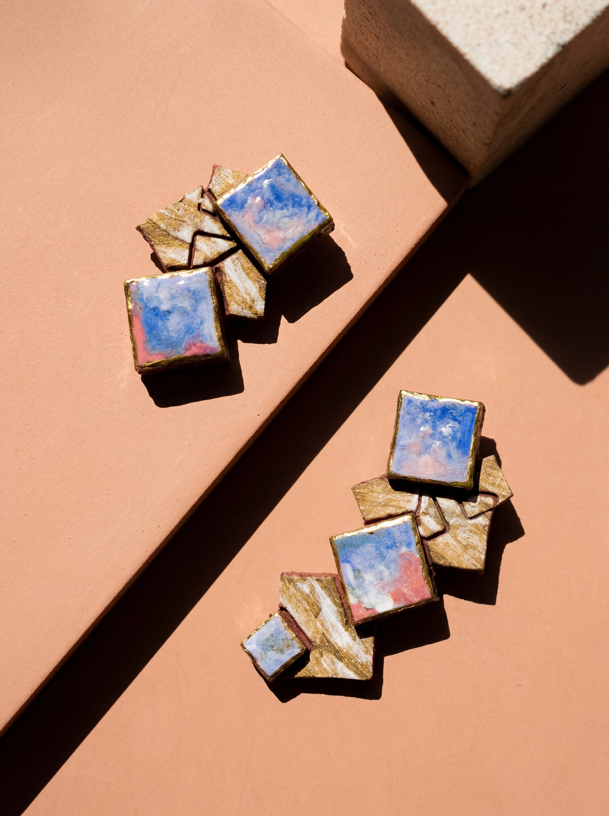 Top-down view of HECROWN 003 geometric ceramic earrings resting on a terracotta clay slab. High-contrast sunlight creates dramatic shadows, emphasizing the layered square fragments and vibrant abstract glazes. Handmade jewelry from Jingdezhen, blending modern architectural form with warm, earthy textures