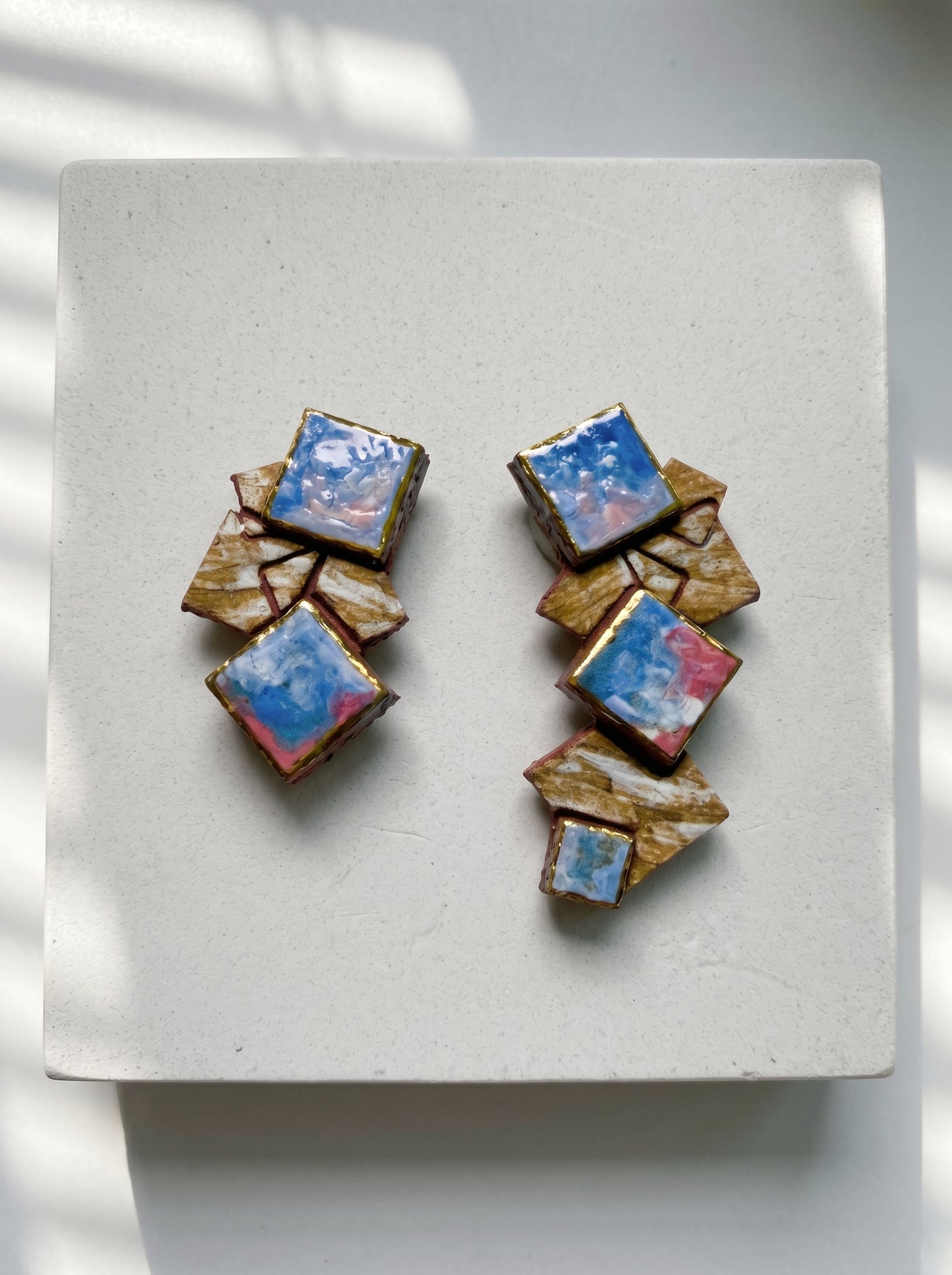 Top-down view of a pair of handmade HECROWN geometric ceramic earrings on a white stone pedestal. Soft diagonal sunlight shadows emphasize the sculptural, asymmetrical square fragments and vibrant blue-pink glazes.
