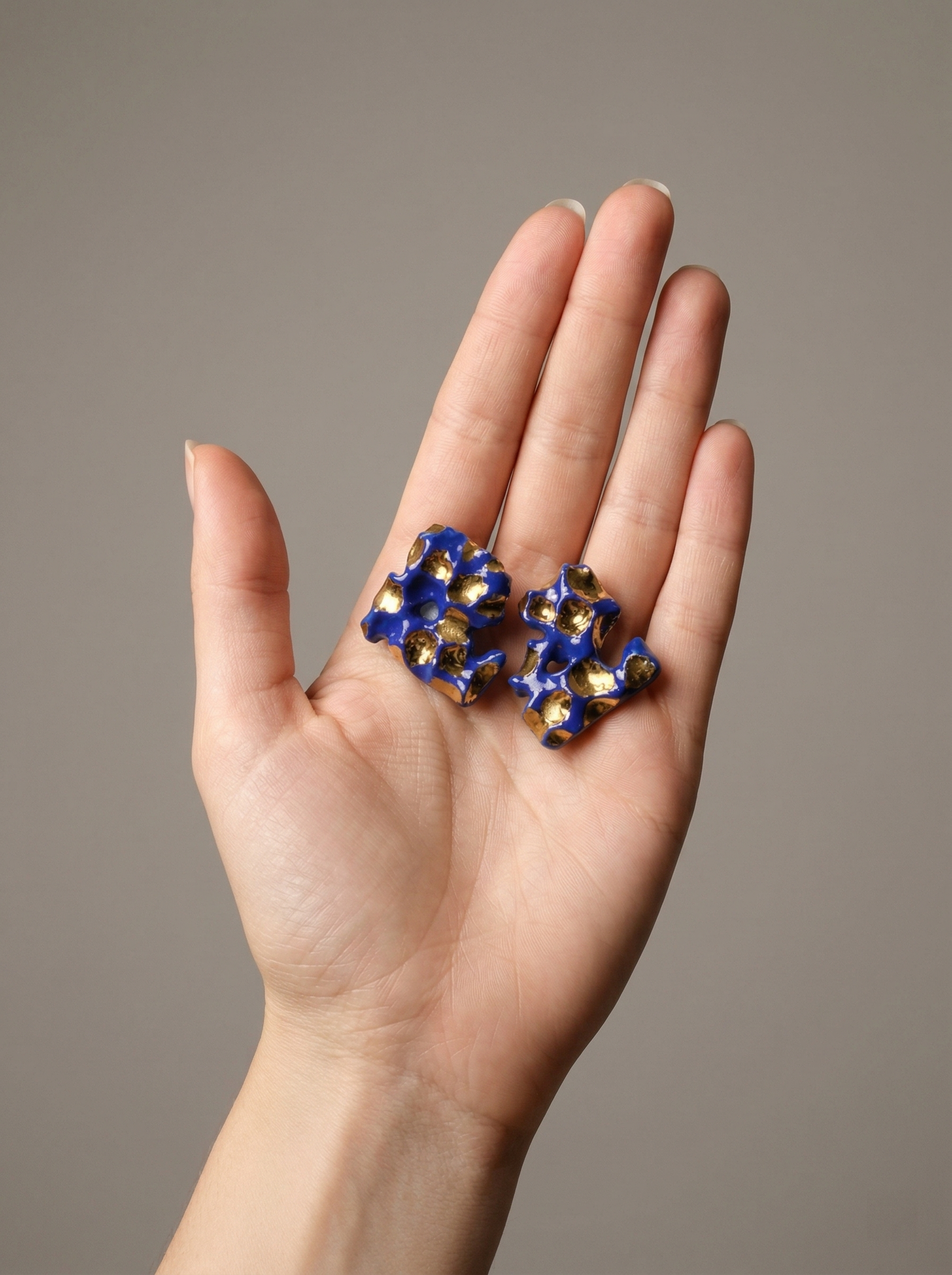 A pair of HECROWN 004 organic ceramic earrings held in a palm for size reference. The deep cobalt blue and liquid gold glaze contrast beautifully with the skin tone. Showcasing the genuine scale and lightweight nature of Jingdezhen artisan jewelry.