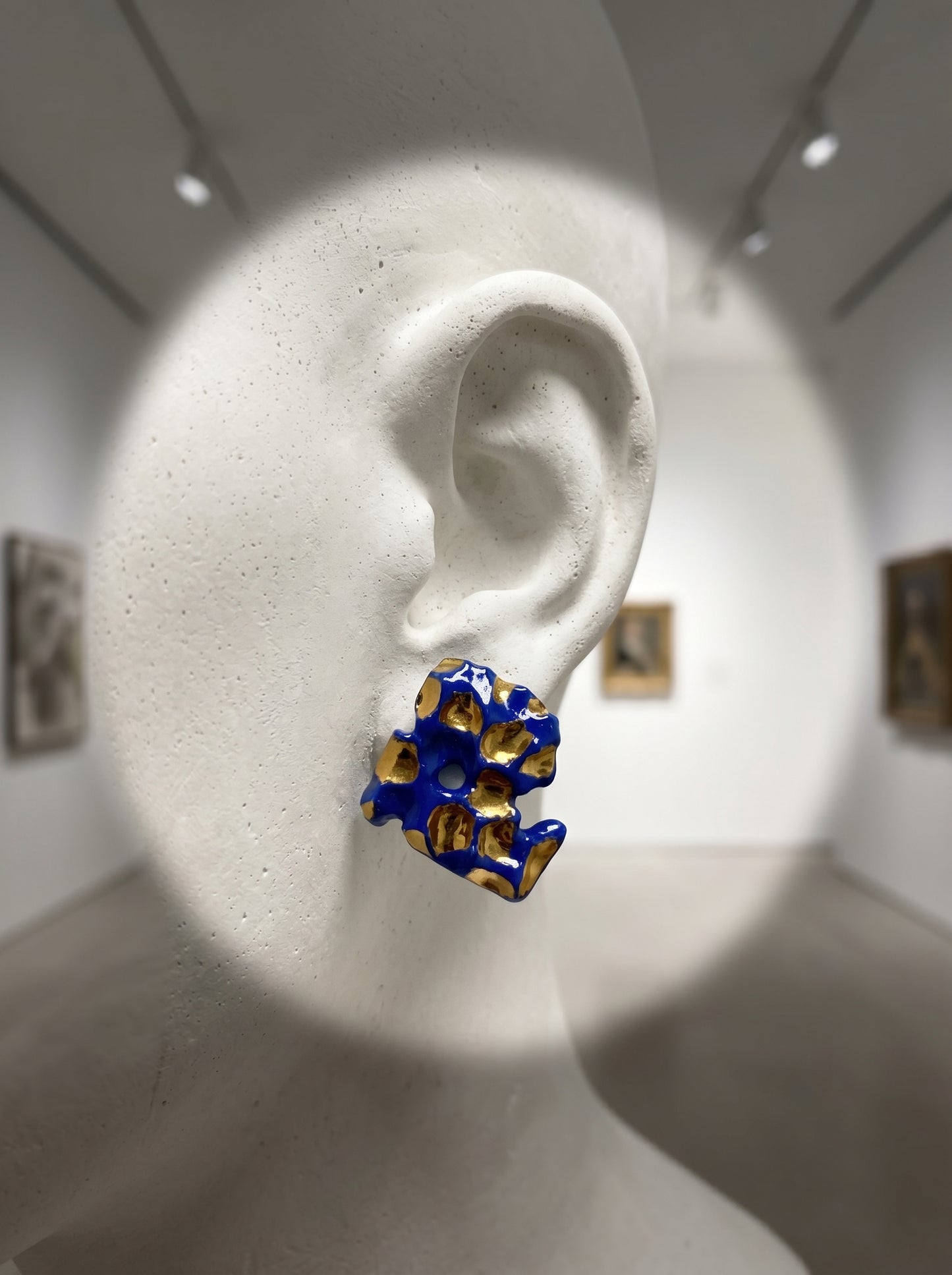 A single HECROWN 004 organic ceramic earring with cobalt blue and gold glaze displayed on a white plaster bust in an art gallery setting. Highlighting the sculptural elegance and artistic soul of handmade Jingdezhen jewelry as a piece of wearable fine art.