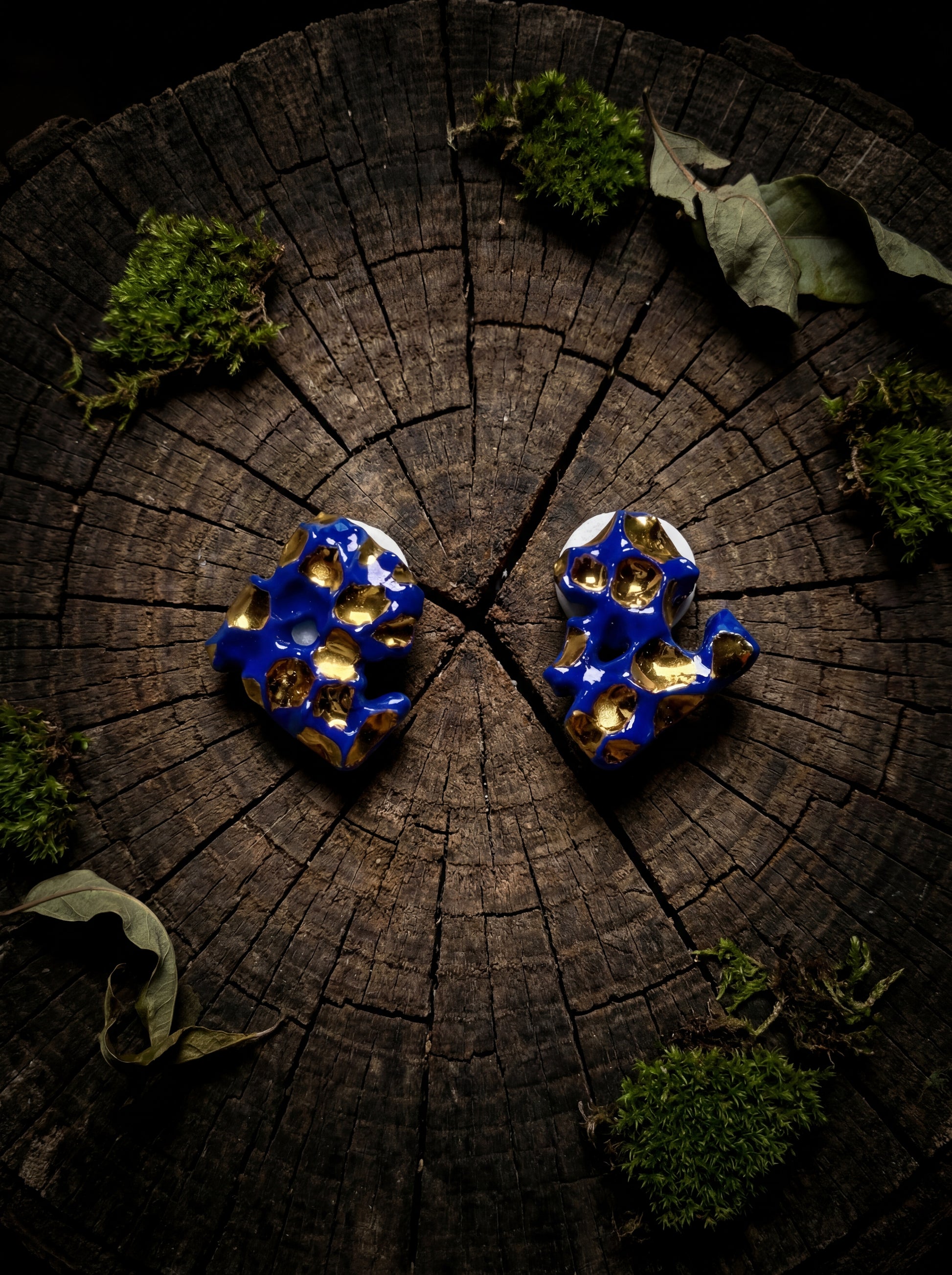 A pair of handmade HECROWN organic ceramic earrings with deep cobalt blue and liquid gold glaze, displayed on a weathered dark wood stump surrounded by green moss and dried leaves. Highlighting the fusion of ancient Jingdezhen craftsmanship and raw natural elements.