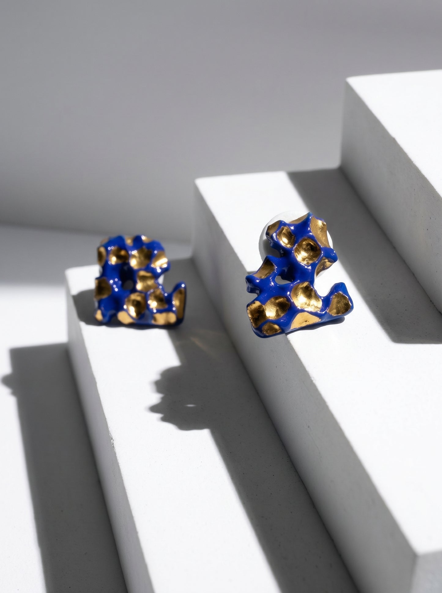 A pair of handmade HECROWN organic ceramic earrings with deep cobalt blue and gold glazes, displayed on a minimalist white geometric staircase. Sharp sunlight creates dramatic architectural shadows, highlighting the sculptural form and Jingdezhen artisan craftsmanship.