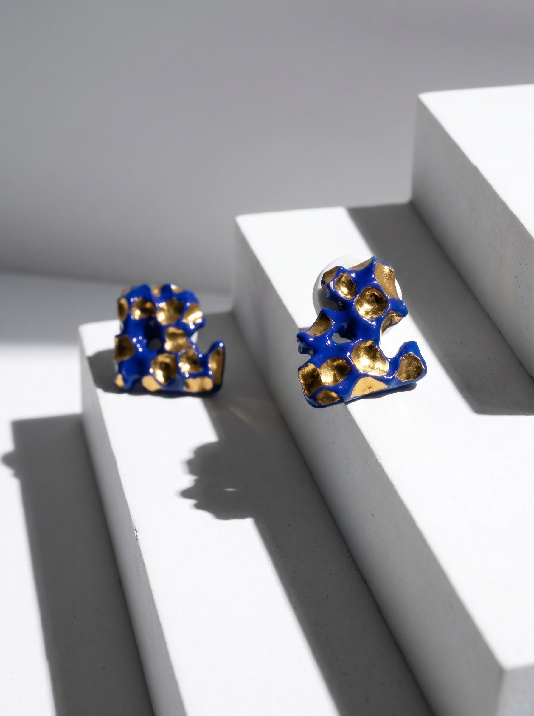 A pair of handmade HECROWN organic ceramic earrings with deep cobalt blue and gold glazes, displayed on a minimalist white geometric staircase. Sharp sunlight creates dramatic architectural shadows, highlighting the sculptural form and Jingdezhen artisan craftsmanship.