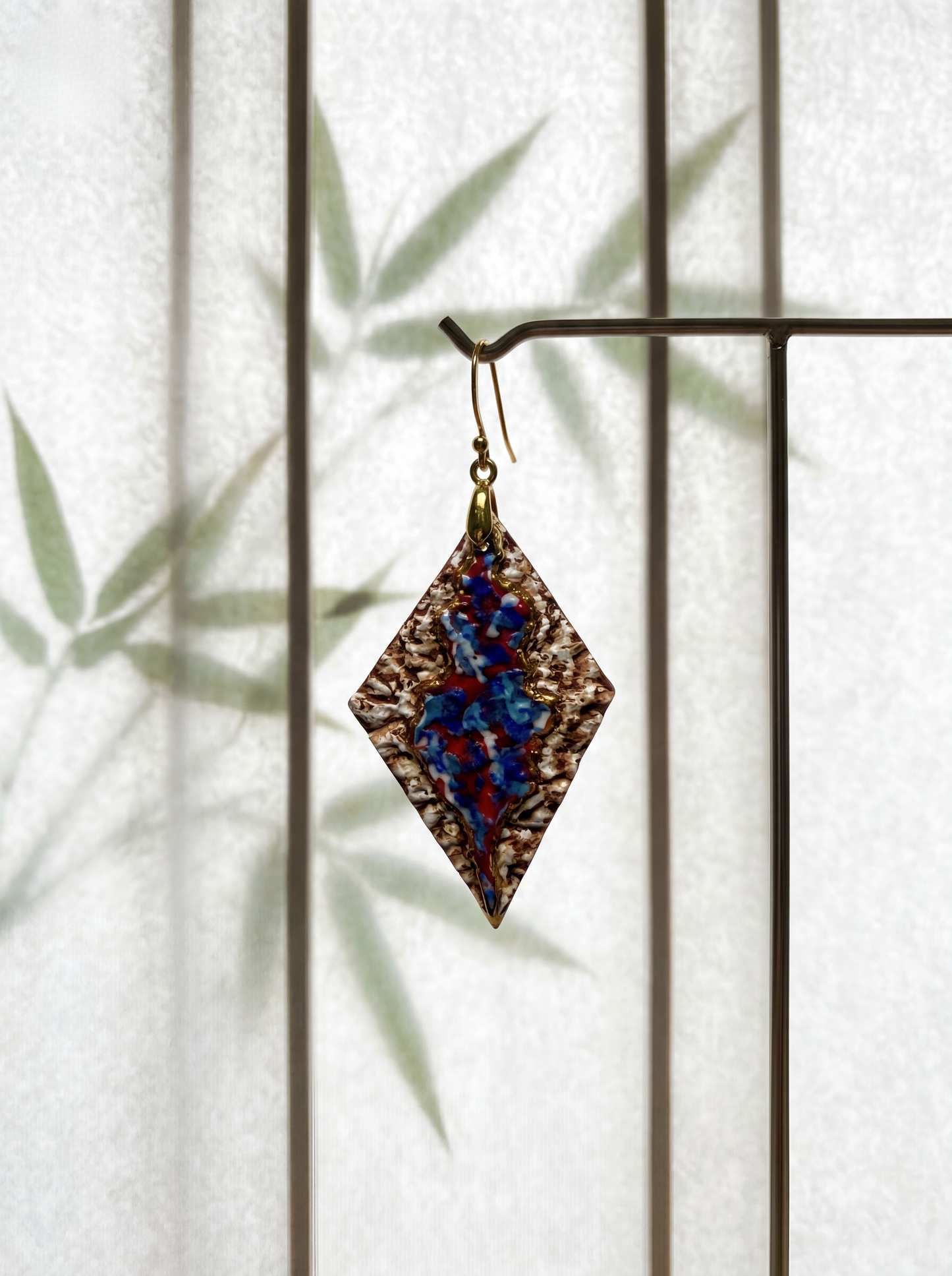 A single HECROWN 005 rhombus ceramic earring suspended on a minimalist metal stand against a translucent screen with bamboo leaf shadows. Highlighting the fusion of rugged handcrafted textures and serene oriental aesthetics from Jingdezhen.