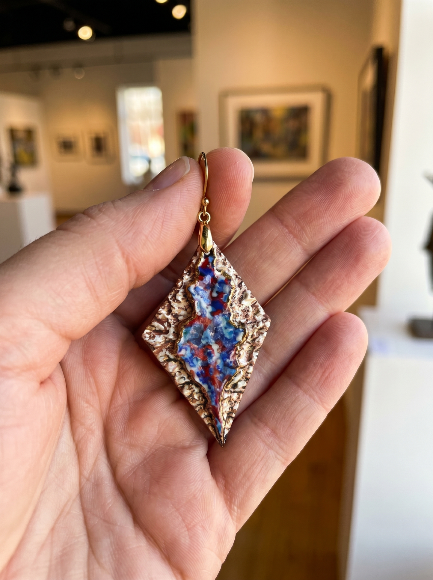 A single HECROWN 005 rhombus ceramic earring held in a palm for scale, set against a blurred art gallery background. The vibrant blue and red flowing glazes and textured bronze surface are clearly visible, showcasing the sincere handcrafted quality from Jingdezhen.