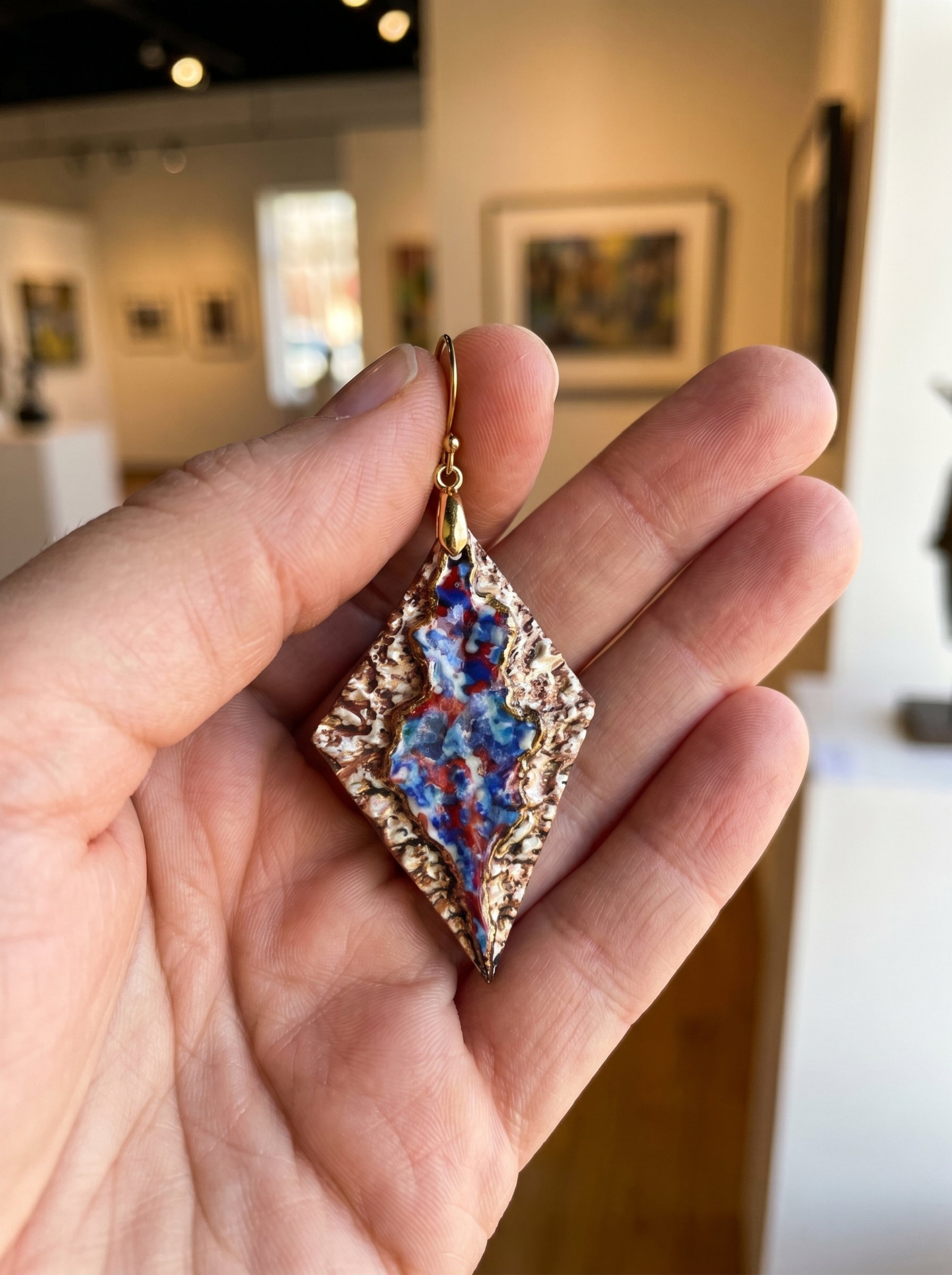 A single HECROWN 005 rhombus ceramic earring held in a palm for scale, set against a blurred art gallery background. The vibrant blue and red flowing glazes and textured bronze surface are clearly visible, showcasing the sincere handcrafted quality from Jingdezhen.