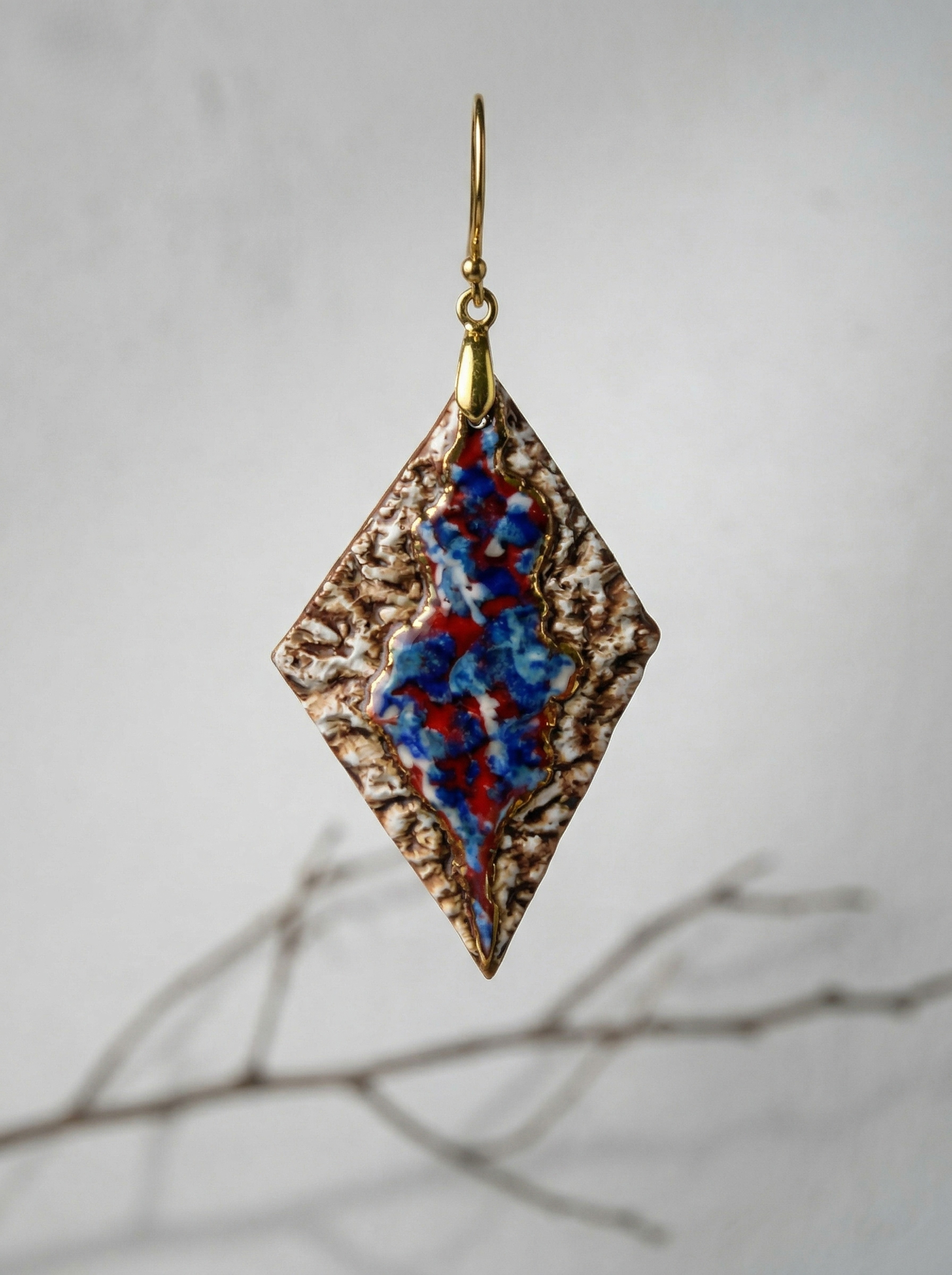 A single HECROWN 005 rhombus ceramic earring suspended in front of a soft grey background with a blurred dry branch. Highlighting the intricate bronze-like texture and vibrant flowing glaze, representing the sincere handcrafted spirit from Jingdezhen.