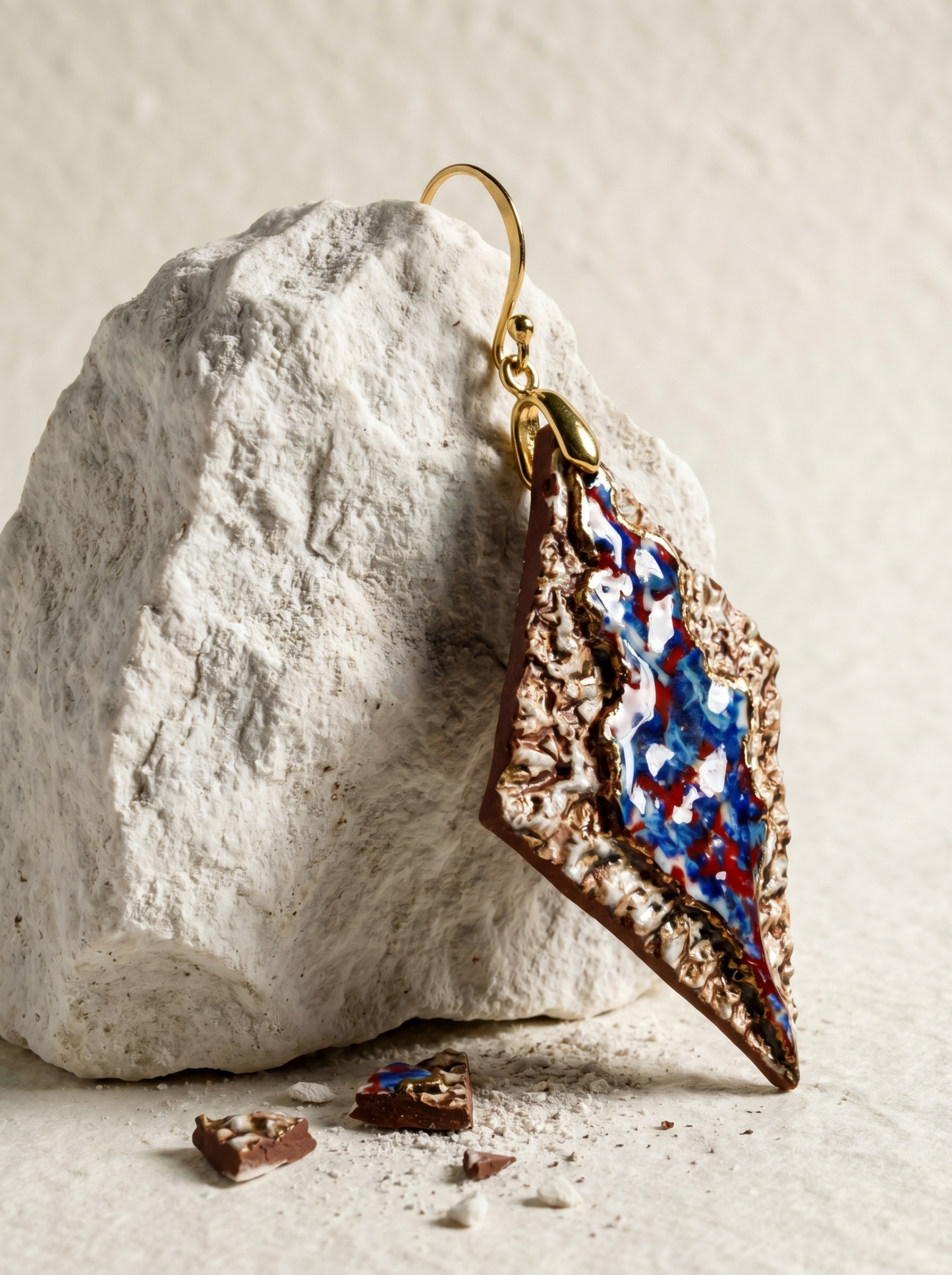A single HECROWN 005 rhombus ceramic earring hanging on a rough white rock. The earthy bronze texture and vibrant blue-red glaze are contrasted with ceramic shards and dust on the ground, showcasing the sincere handcrafted process from Jingdezhen.
