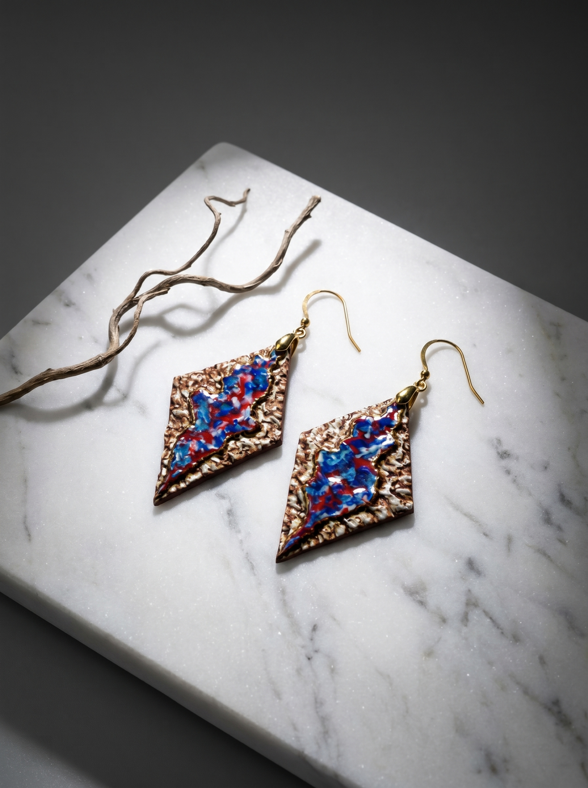 A pair of handmade rhombus ceramic earrings by HECROWN, featuring a textured earthy bronze surface and a vibrant blue-red glazed center. Displayed on a white marble slab next to a dry branch under focused lighting, showcasing the intricate handcrafted details from Jingdezhen.