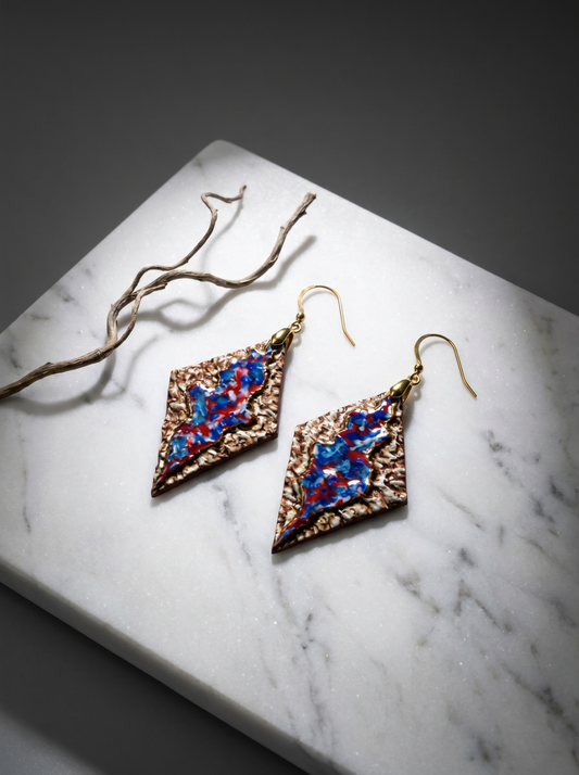 A pair of handmade rhombus ceramic earrings by HECROWN, featuring a textured earthy bronze surface and a vibrant blue-red glazed center. Displayed on a white marble slab next to a dry branch under focused lighting, showcasing the intricate handcrafted details from Jingdezhen.