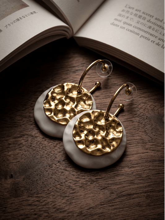 A pair of HECROWN 006 handmade ceramic earrings featuring hammered gold plates on wavy white porcelain bases, displayed on a wooden surface next to an open book.