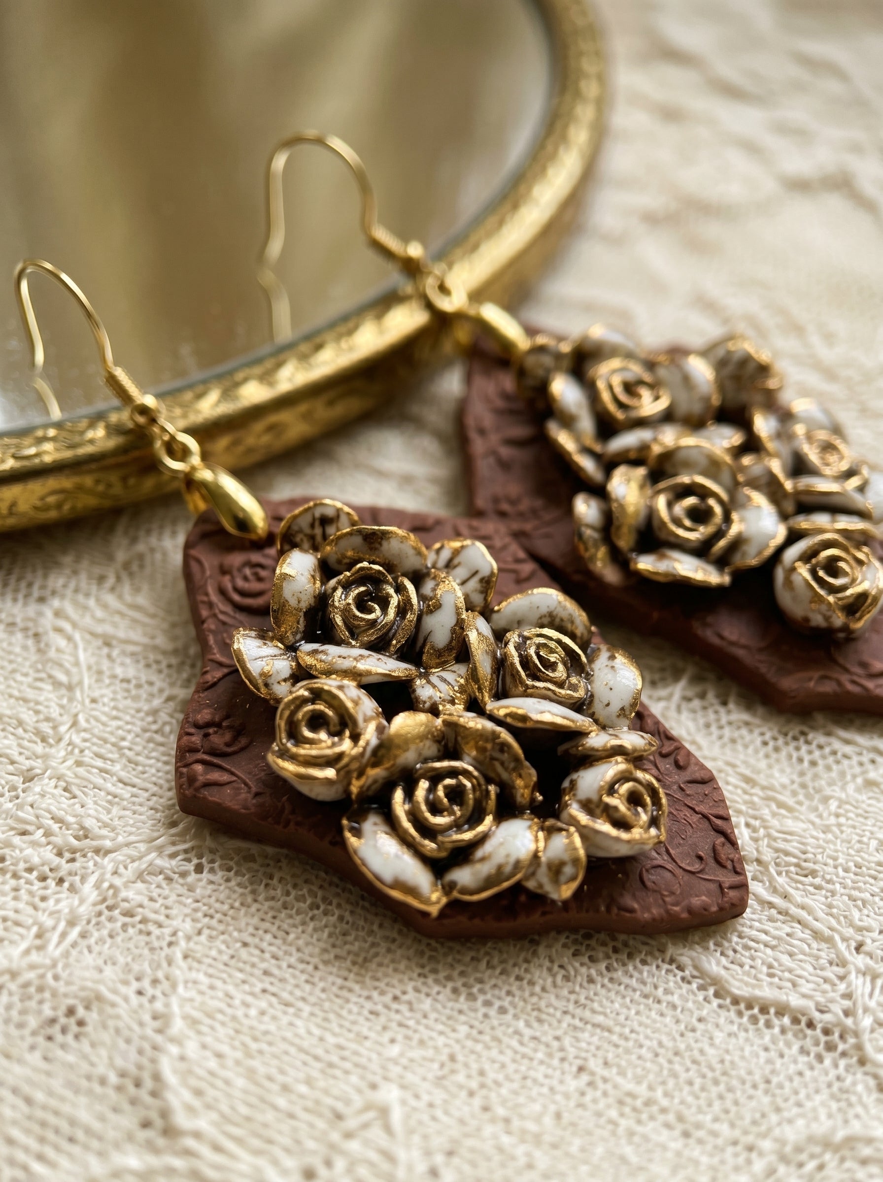 Close-up of HECROWN 007 ceramic earrings on a lace cloth next to a vintage gold mirror, representing a warm, realistic, and nostalgic handcrafted aesthetic.