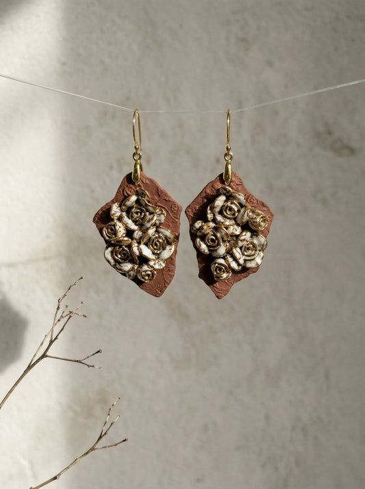 A pair of HECROWN 007 Ruined Rose ceramic earrings featuring white porcelain roses on terracotta-colored textured bases, suspended against a bamboo shadow backdrop.