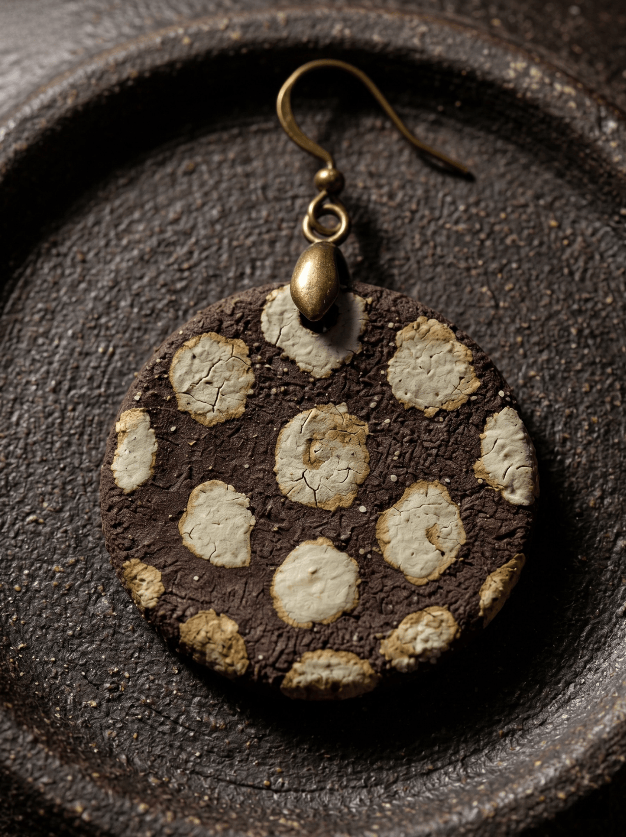 Extreme macro shot of a HECROWN rustic ceramic drop earring resting on a dark textured plate, highlighting the intricate ice crackle glaze. Sincere wabi-sabi artisan gift.