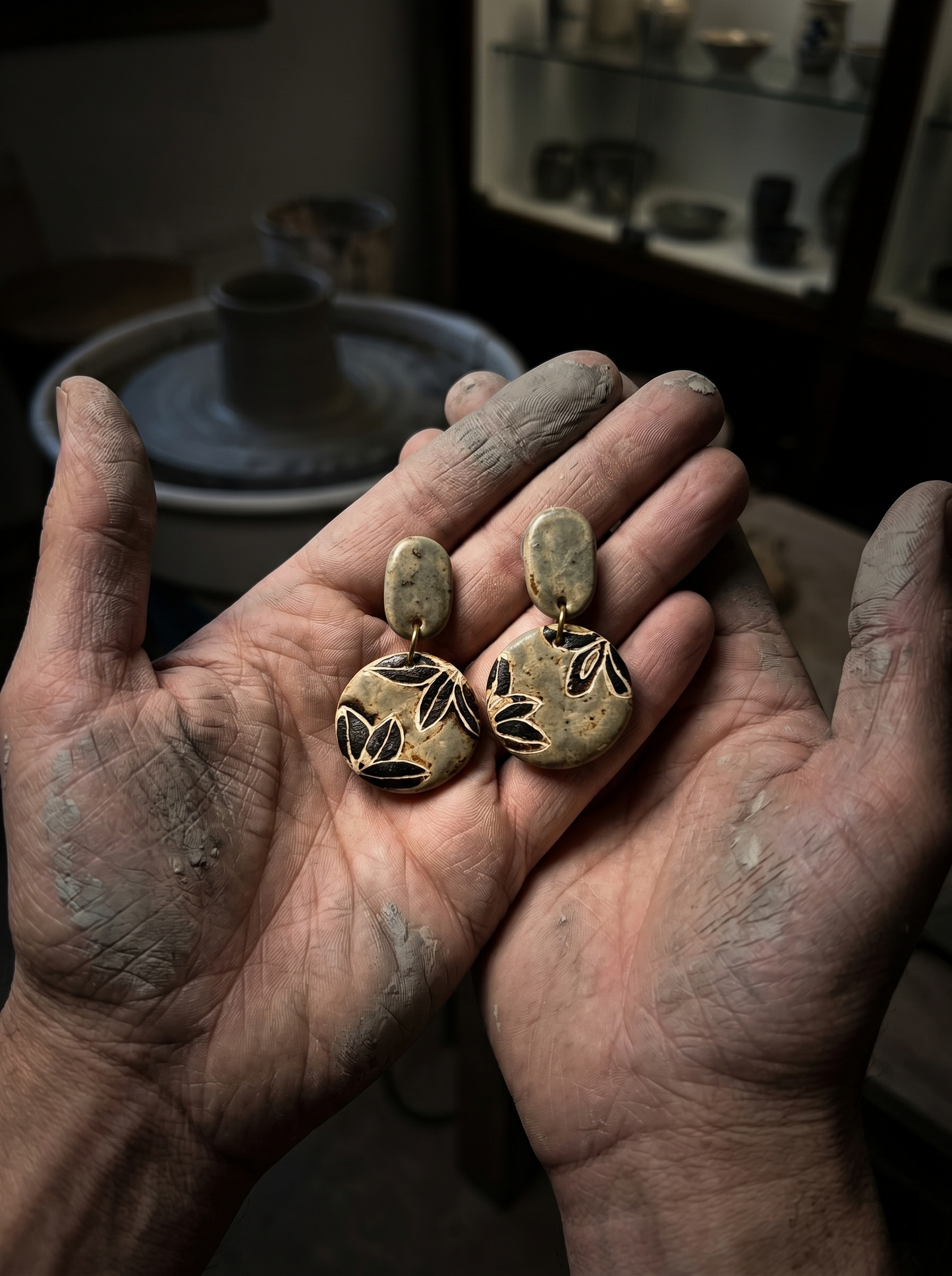 HECROWN celadon botanical ceramic earrings held in the clay-covered hands of an artisan in a Jingdezhen pottery studio. The ultimate proof of sincere wabi-sabi handmade jewelry.