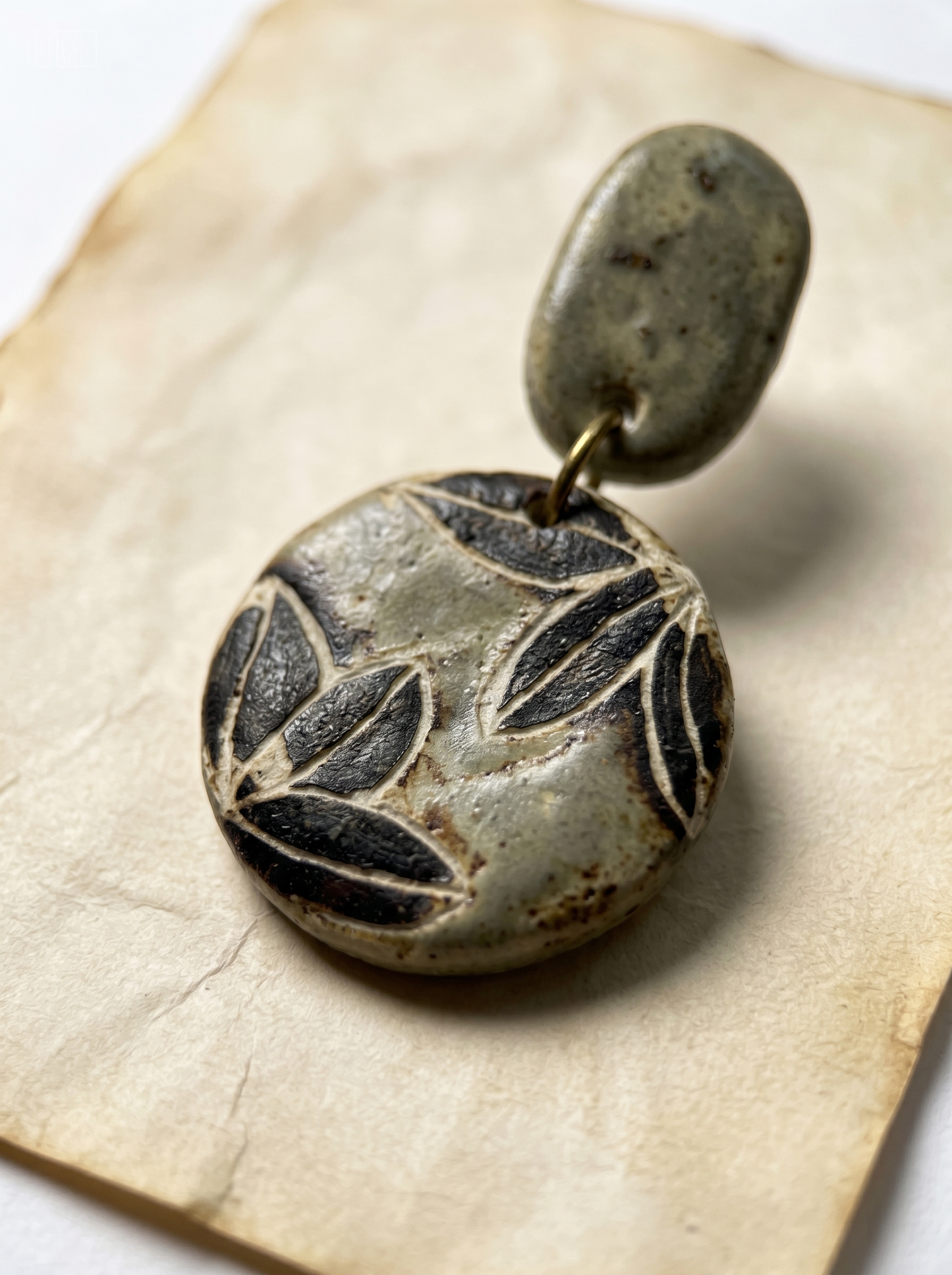 Extreme macro close-up of a HECROWN sage green ceramic earring on vintage paper, highlighting the deep carved leaf texture and rustic glaze. Sincere wabi-sabi handmade jewelry.
