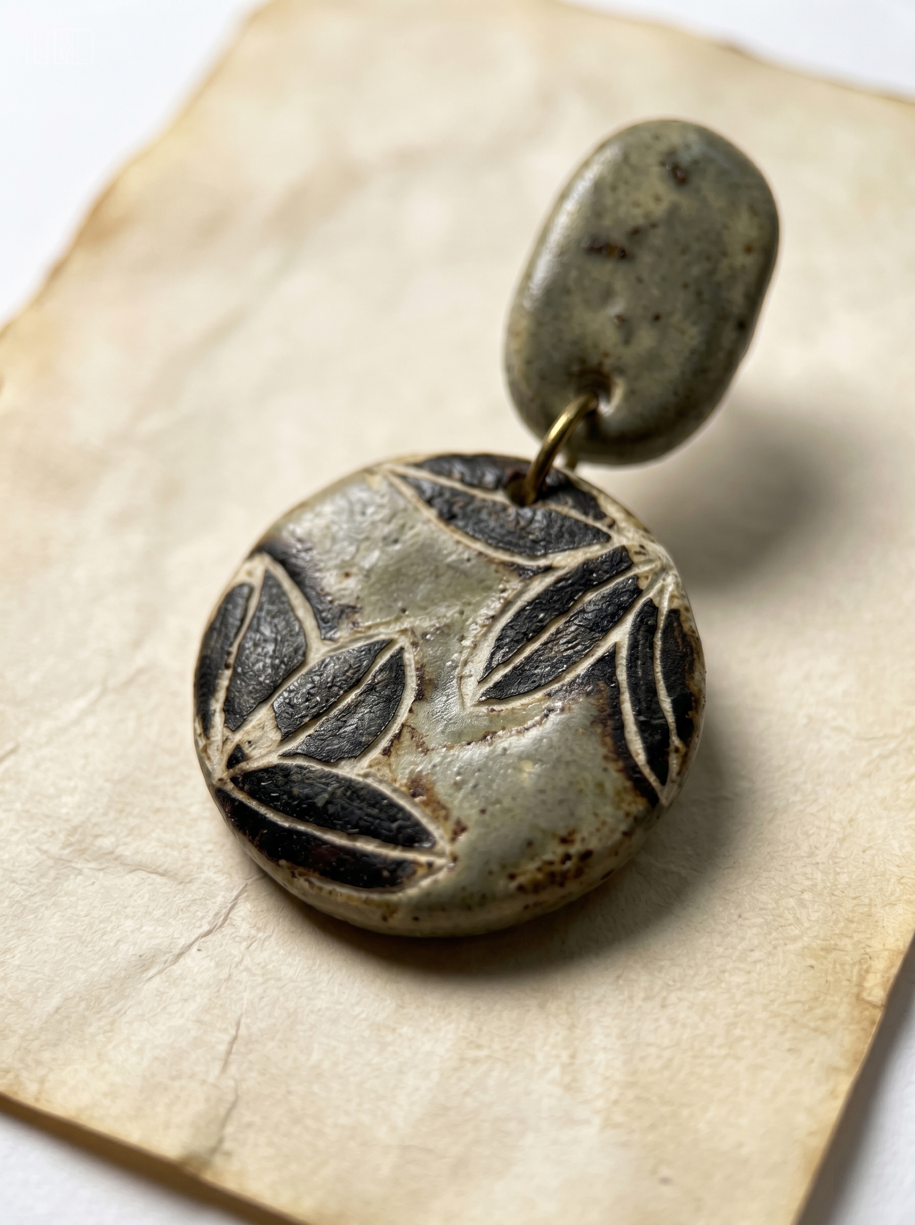 Extreme macro close-up of a HECROWN sage green ceramic earring on vintage paper, highlighting the deep carved leaf texture and rustic glaze. Sincere wabi-sabi handmade jewelry.