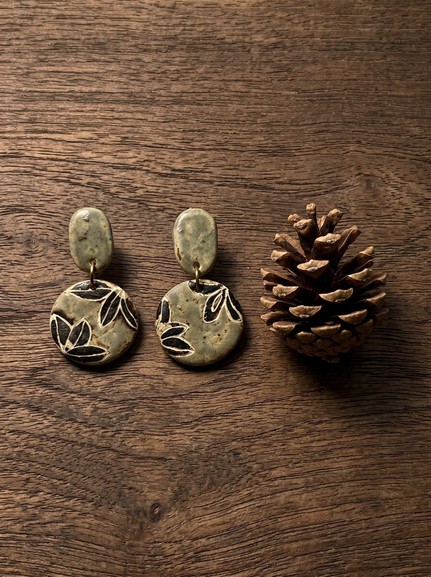 A pair of HECROWN carved botanical ceramic earrings resting on dark wood next to a pinecone. Sincere, nature-inspired handcrafted jewelry from Jingdezhen.