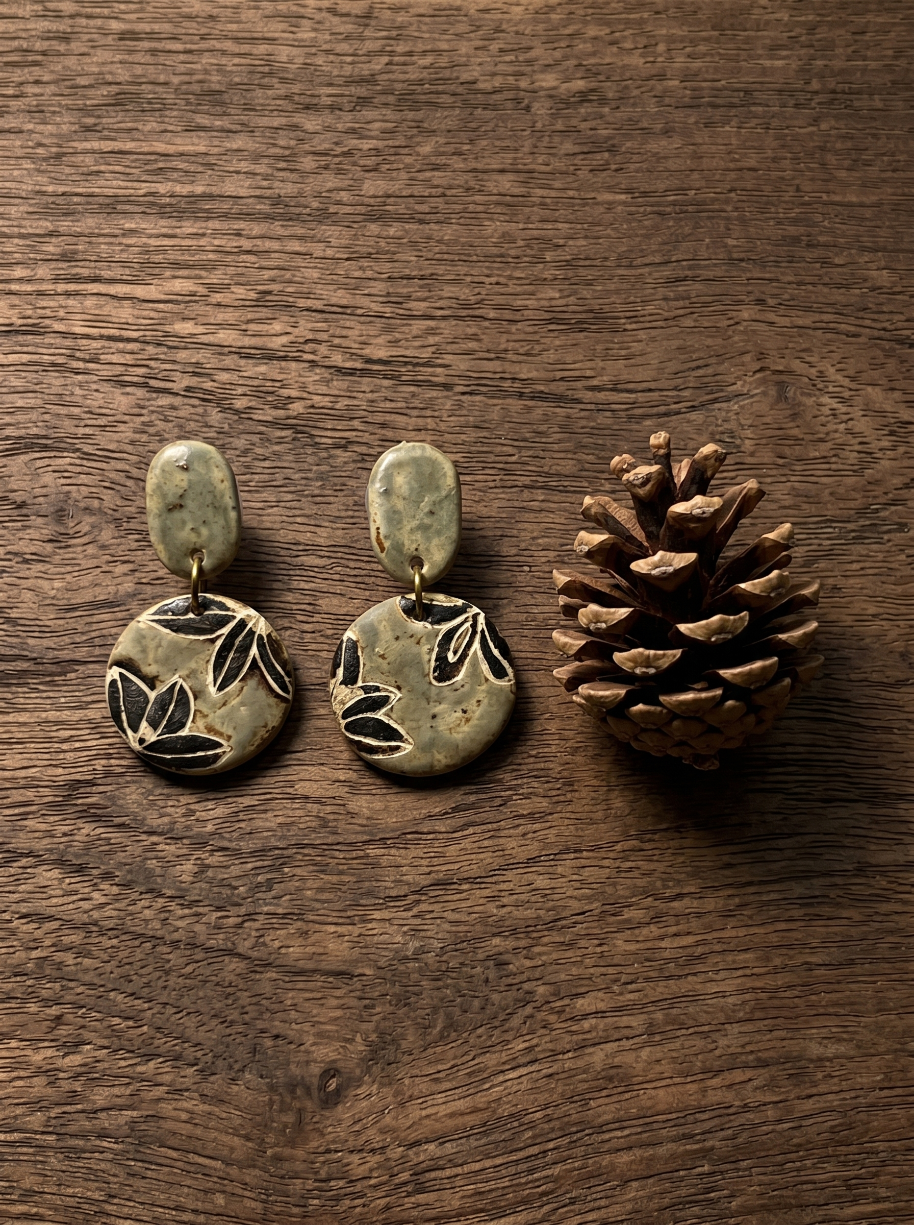 A pair of HECROWN carved botanical ceramic earrings resting on dark wood next to a pinecone. Sincere, nature-inspired handcrafted jewelry from Jingdezhen.