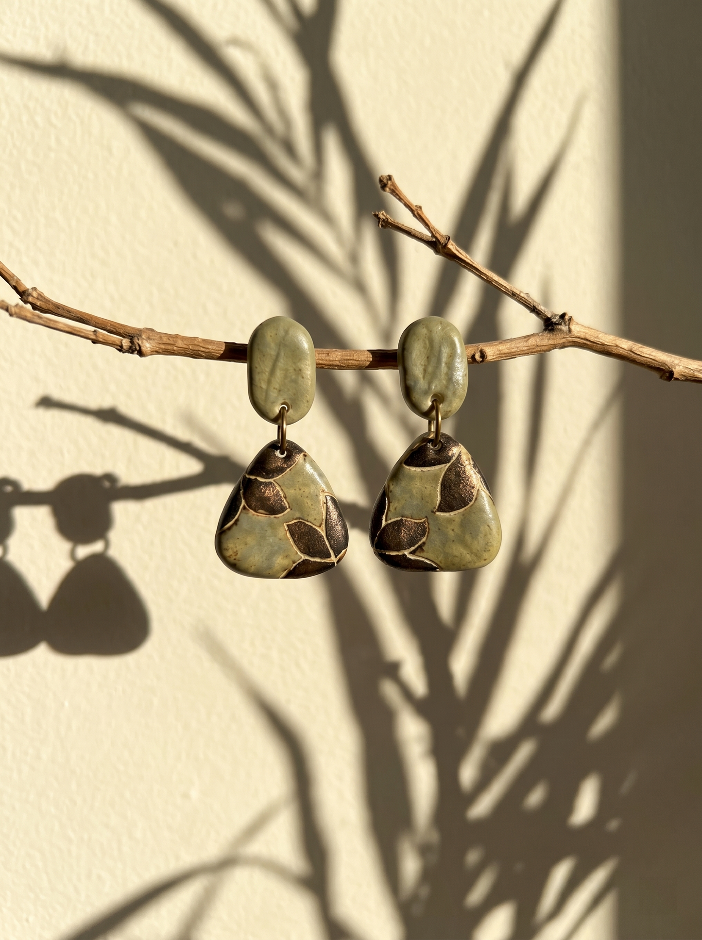 A pair of HECROWN celadon botanical ceramic triangle earrings hanging from a dry branch with leaf shadows. Nature-inspired handmade ceramic art and a unique anniversary gift.