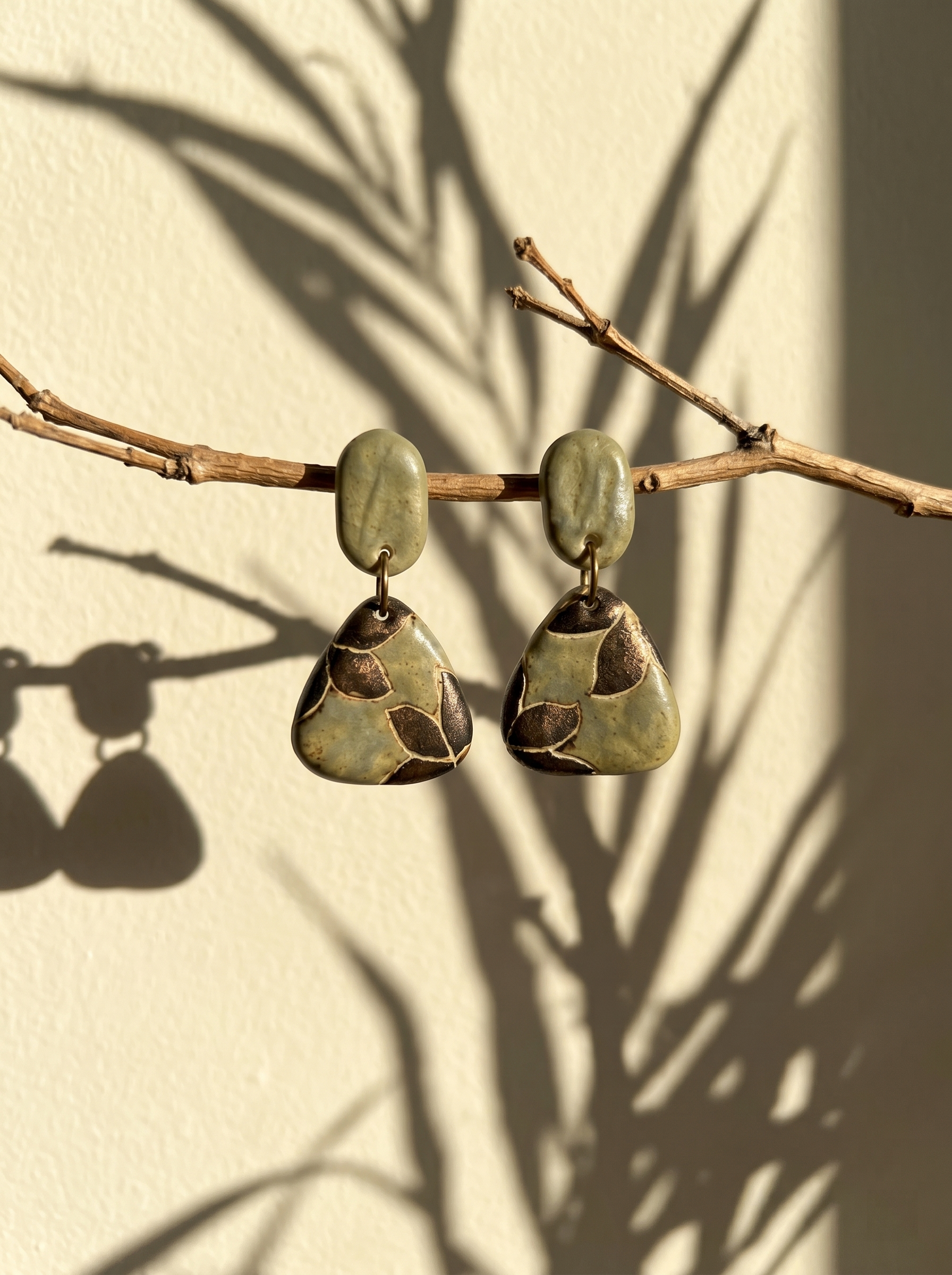 A pair of HECROWN celadon botanical ceramic triangle earrings hanging from a dry branch with leaf shadows. Nature-inspired handmade ceramic art and a unique anniversary gift.
