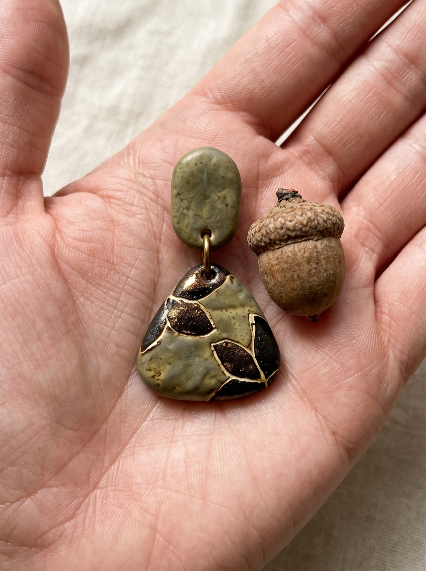 HECROWN carved botanical ceramic earring held in a hand next to a natural acorn for size reference. Highlighting the organic, sincere aesthetic of handcrafted jewelry.