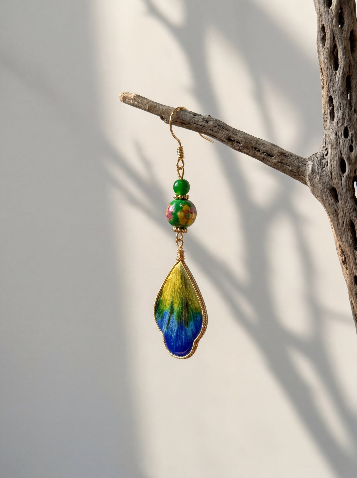 A single HECROWN ceramic and silk feather earring in blue and yellow, hanging from a natural branch. Unique Jingdezhen handmade jewelry and wearable art.