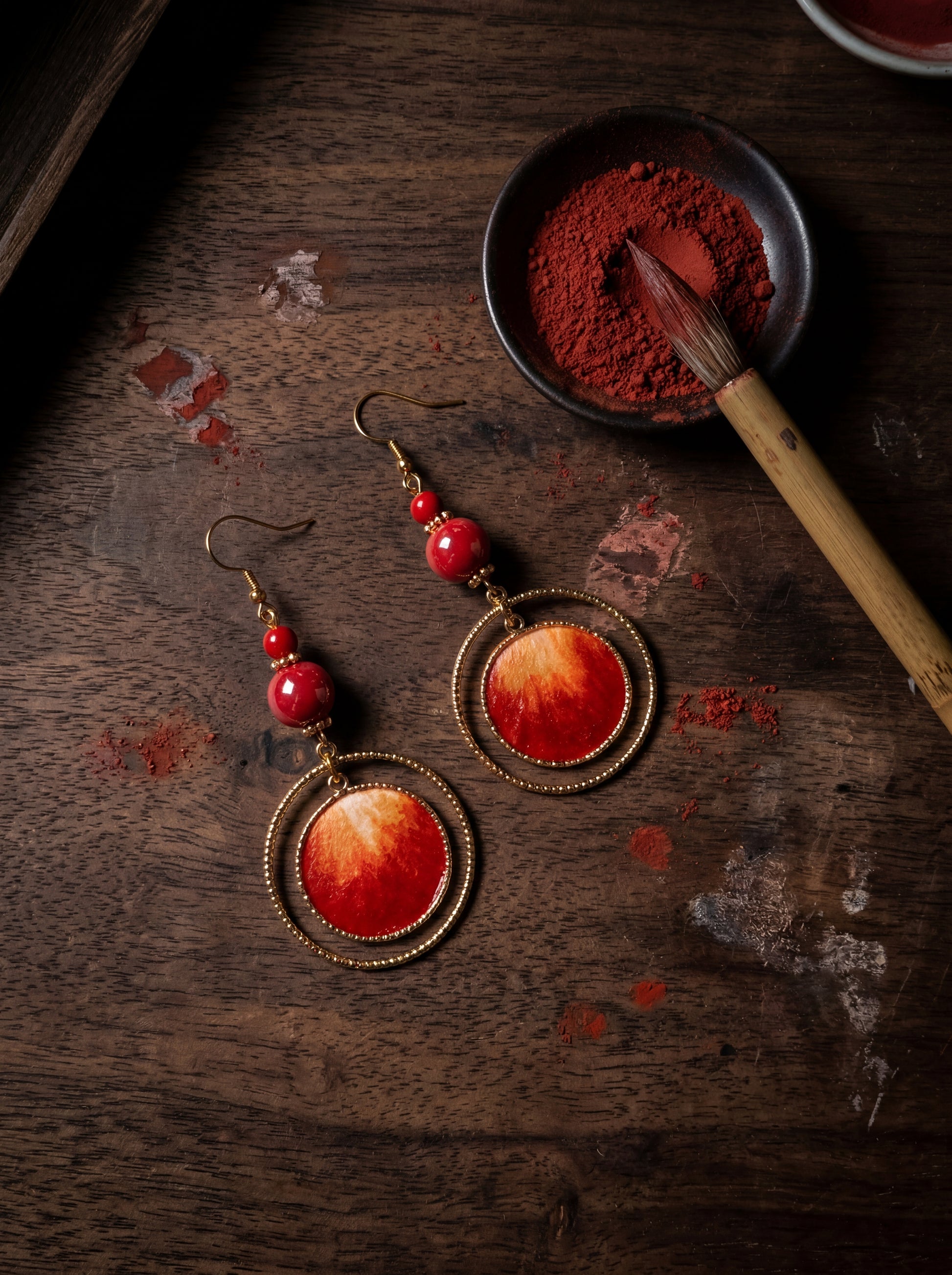 Macro close-up of HECROWN red silk earrings on a workbench with mineral pigments and an artisan brush. Authentic Jingdezhen craftsmanship proof.