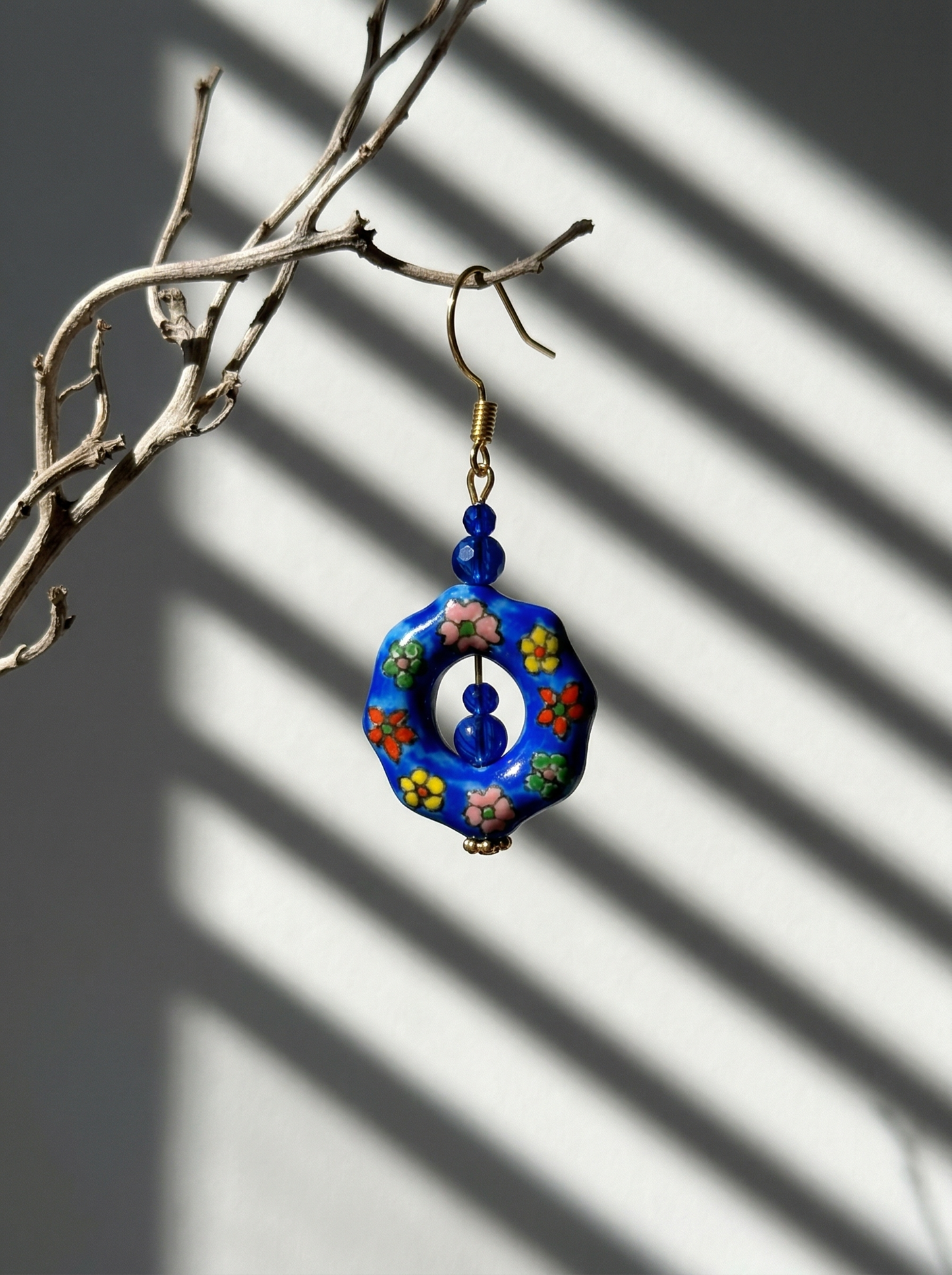 A single HECROWN cobalt blue ceramic hoop earring with hand-painted colorful flowers, hanging from a branch. Unique Jingdezhen handmade jewelry.