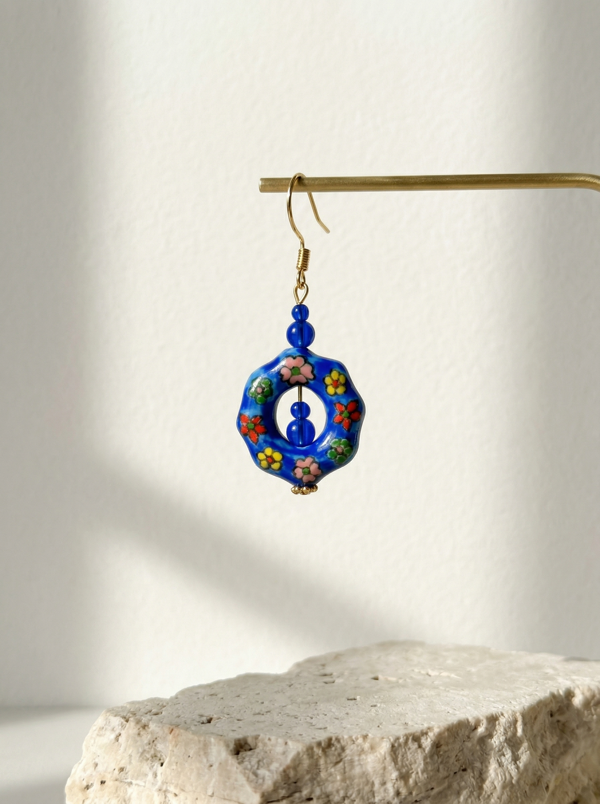 HECROWN hand-painted blue ceramic earring on a gold T-stand against a clean off-white background. Authentic handcrafted porcelain jewelry.