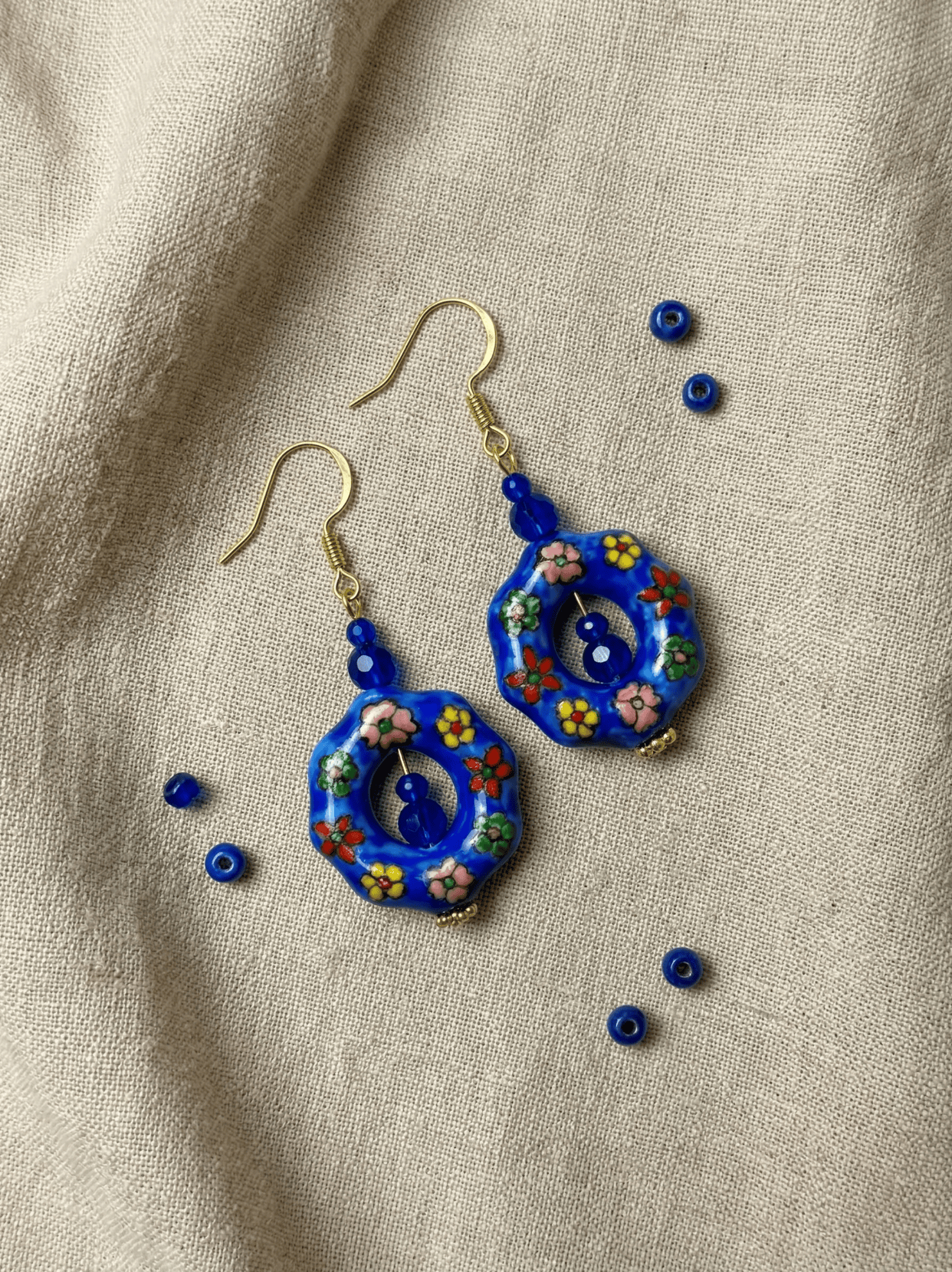 HECROWN hand-painted earrings on light linen with scattered blue glass beads. A warm lifestyle shot of Jingdezhen handmade jewelry.