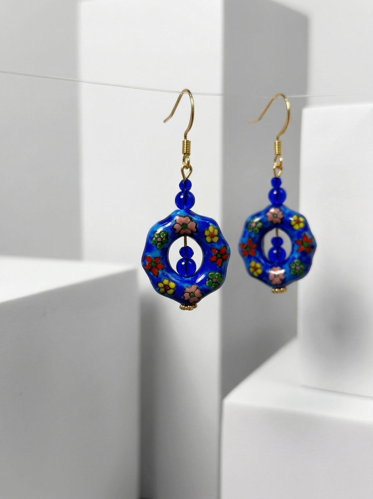 A pair of HECROWN blue floral ceramic earrings suspended on a thin wire between white minimalist blocks. Sincere Jingdezhen ceramic art.