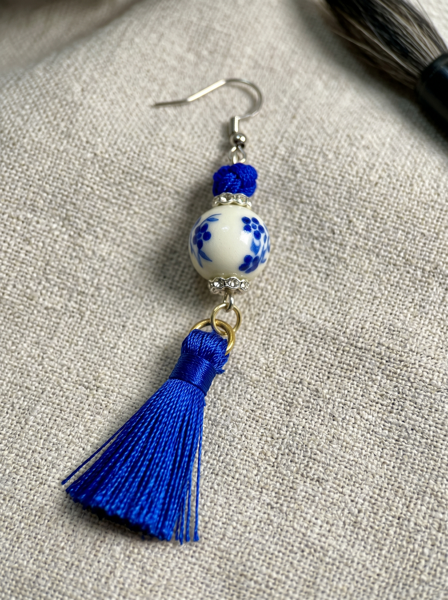Extreme macro close-up of HECROWN earring, focusing on the hand-painted blue flowers and fine silk tassel threads. Sincere handcrafted jewelry.