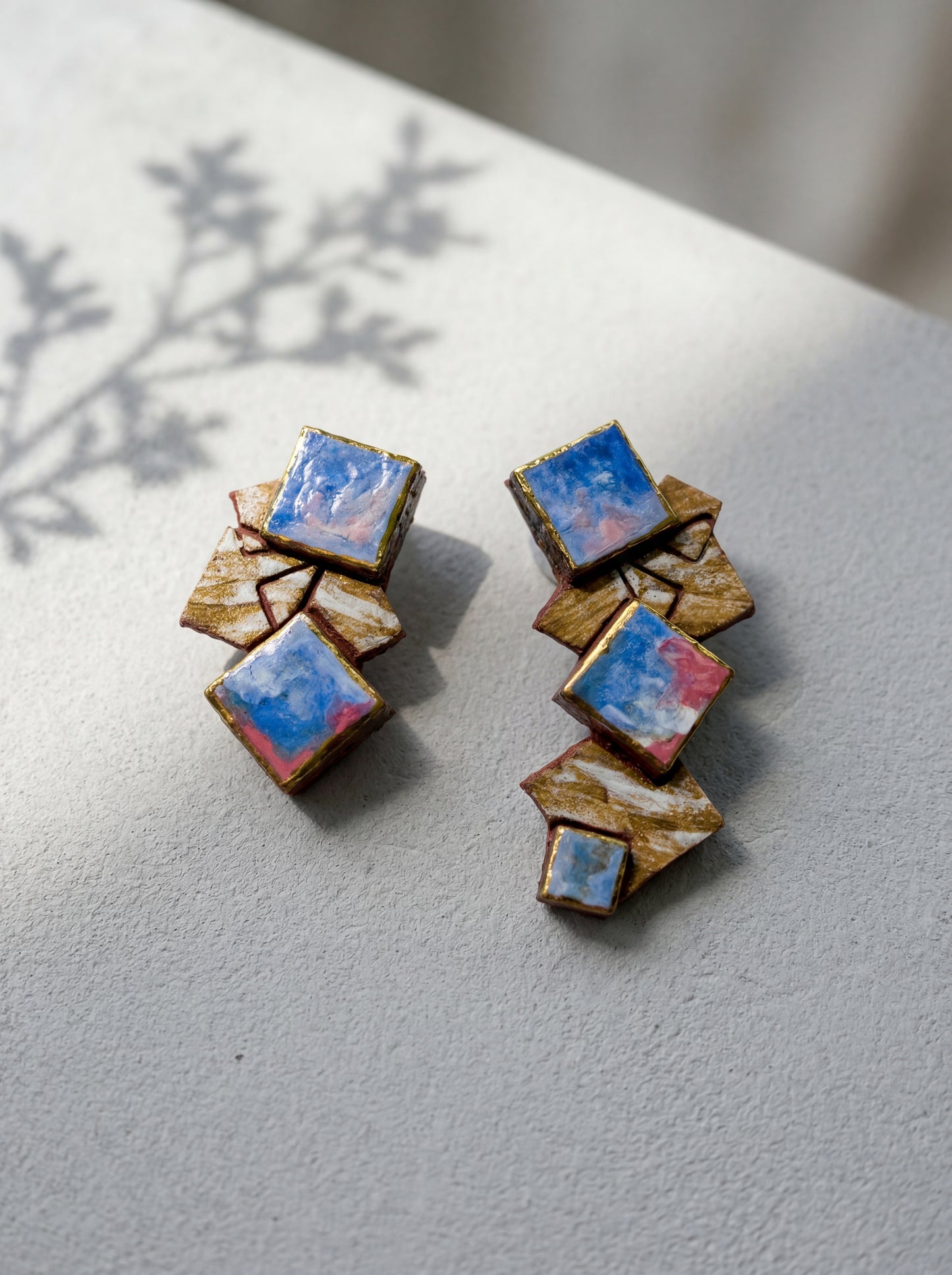 A pair of geometric handmade ceramic stud earrings featuring layered square fragments in a blue and pink abstract glaze, resting on a textured light grey surface with botanical shadows. HECROWN contemporary jewelry from Jingdezhen, blending sculptural form with painterly textures.