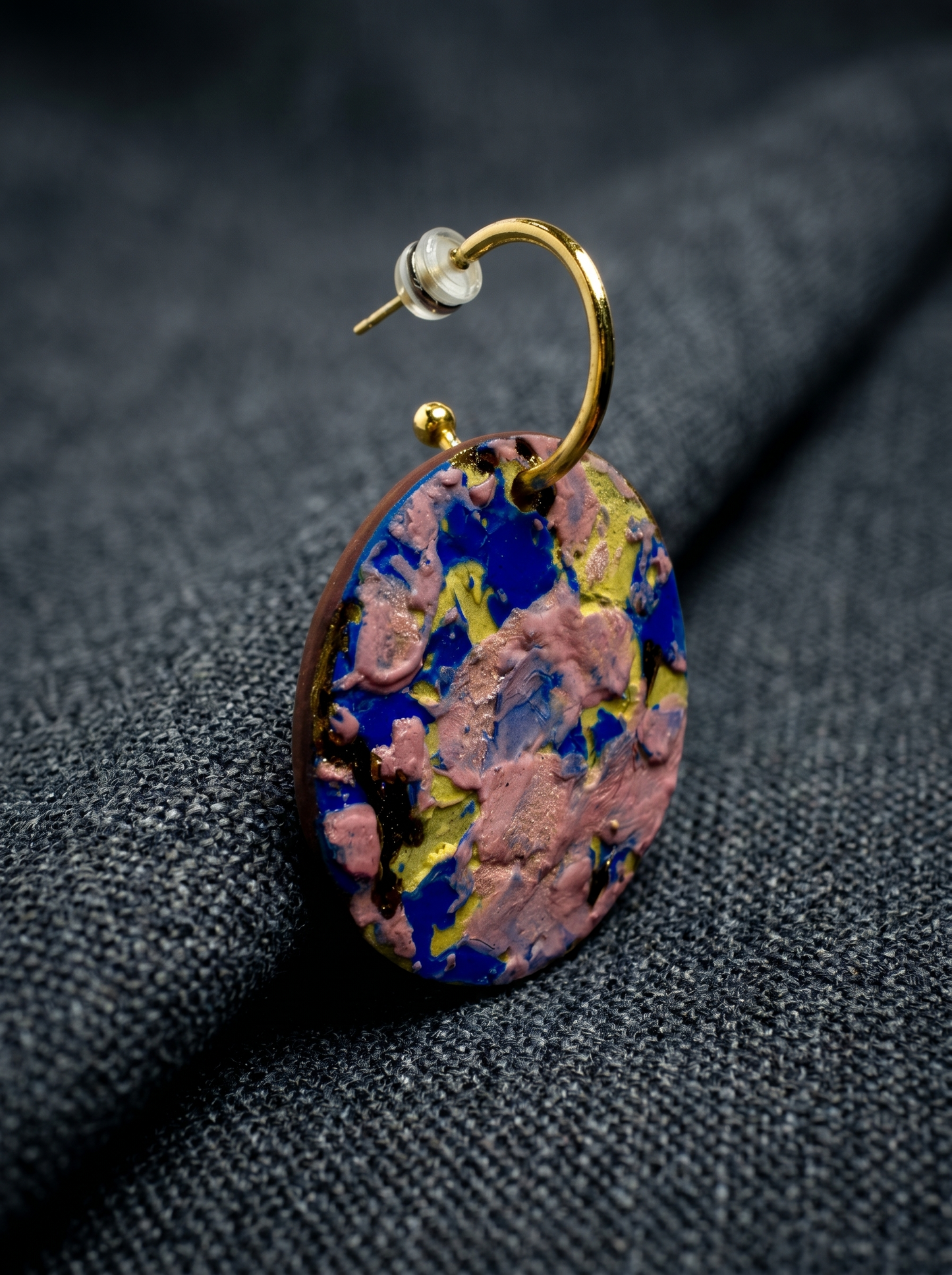 Extreme close-up of a handmade ceramic earring with vibrant pink and blue impasto glaze, resting on a textured dark grey fabric. HECROWN signature collection detail shot, capturing the intricate physical textures and handcrafted soul of Jingdezhen artistry.