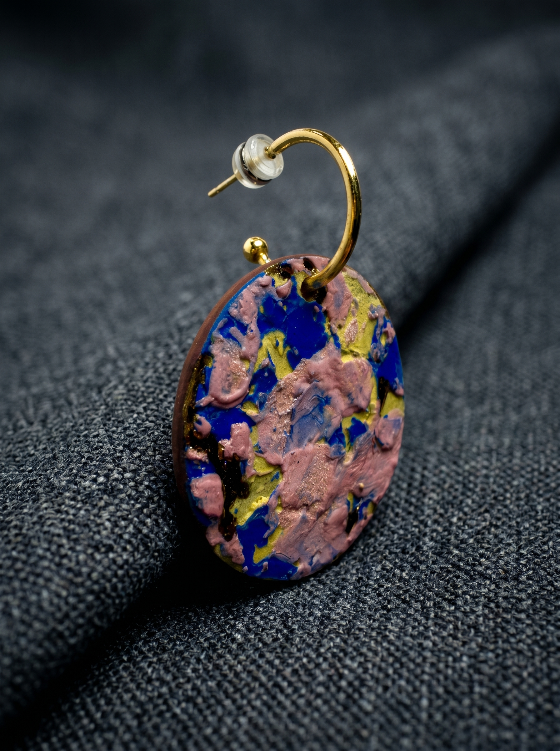 Extreme close-up of a handmade ceramic earring with vibrant pink and blue impasto glaze, resting on a textured dark grey fabric. HECROWN signature collection detail shot, capturing the intricate physical textures and handcrafted soul of Jingdezhen artistry.