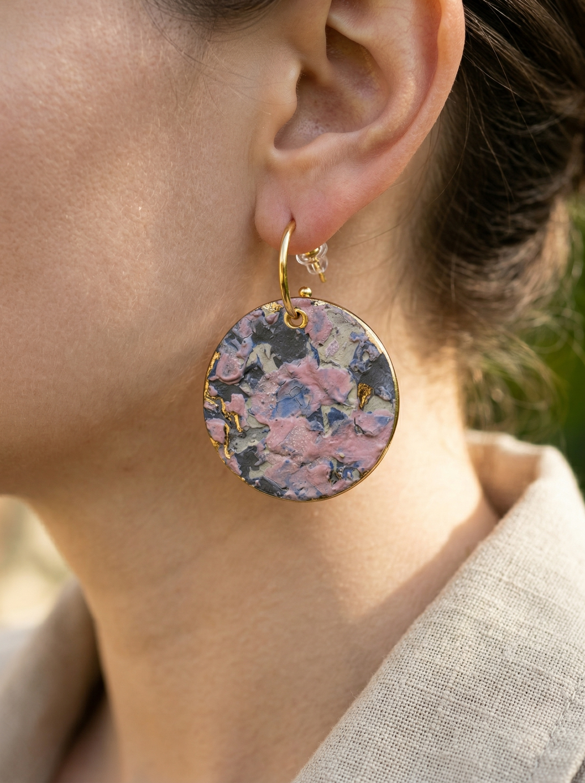 Close-up of a model wearing large round ceramic earrings with vibrant impasto textures in pink and blue, styled with a beige linen shirt. HECROWN handmade artisan jewelry captured in natural outdoor sunlight, emphasizing the organic texture and wearable art aesthetic from Jingdezhen