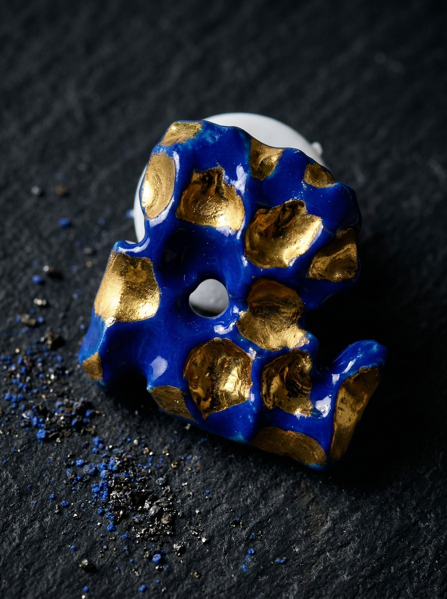 Macro shot of a HECROWN organic ceramic earring featuring deep cobalt blue and liquid gold glazes, resting on a dark textured surface with scattered pigment powders. Highlighting the sculptural details and artistic sincerity of Jingdezhen handmade jewelry.