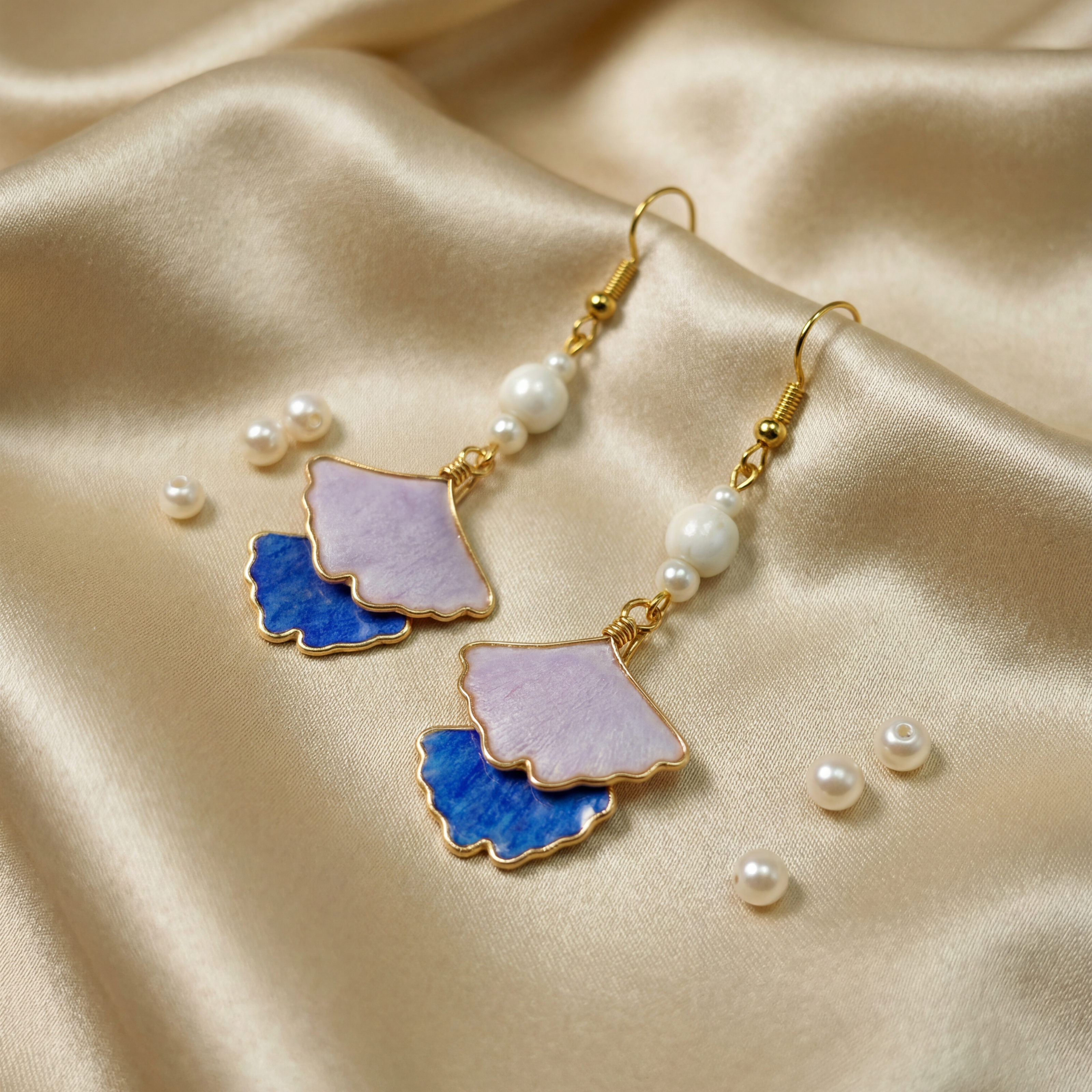 Close-up of elegant dangle earrings featuring double-layered ceramic petals in pastel purple and cobalt blue with gold edges and pearl accents, resting on smooth champagne silk. HECROWN artisan jewelry handcrafted in Jingdezhen, blending Eastern heritage with a romantic, feminine aesthetic.