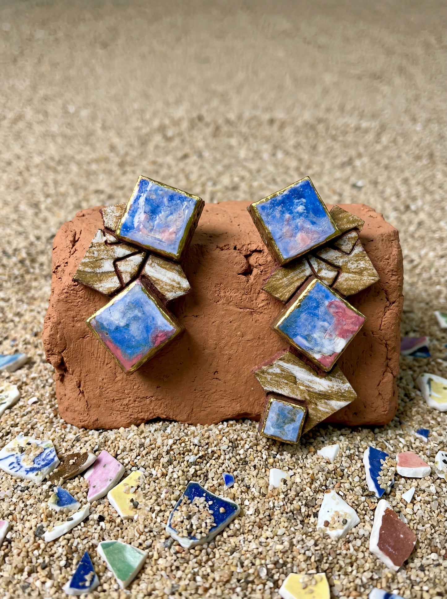 Handmade geometric ceramic earrings displayed on a rustic red brick, surrounded by scattered porcelain shards and fine sand. HECROWN contemporary jewelry from Jingdezhen, blending modern abstract patterns with the raw beauty of traditional kiln fragments.