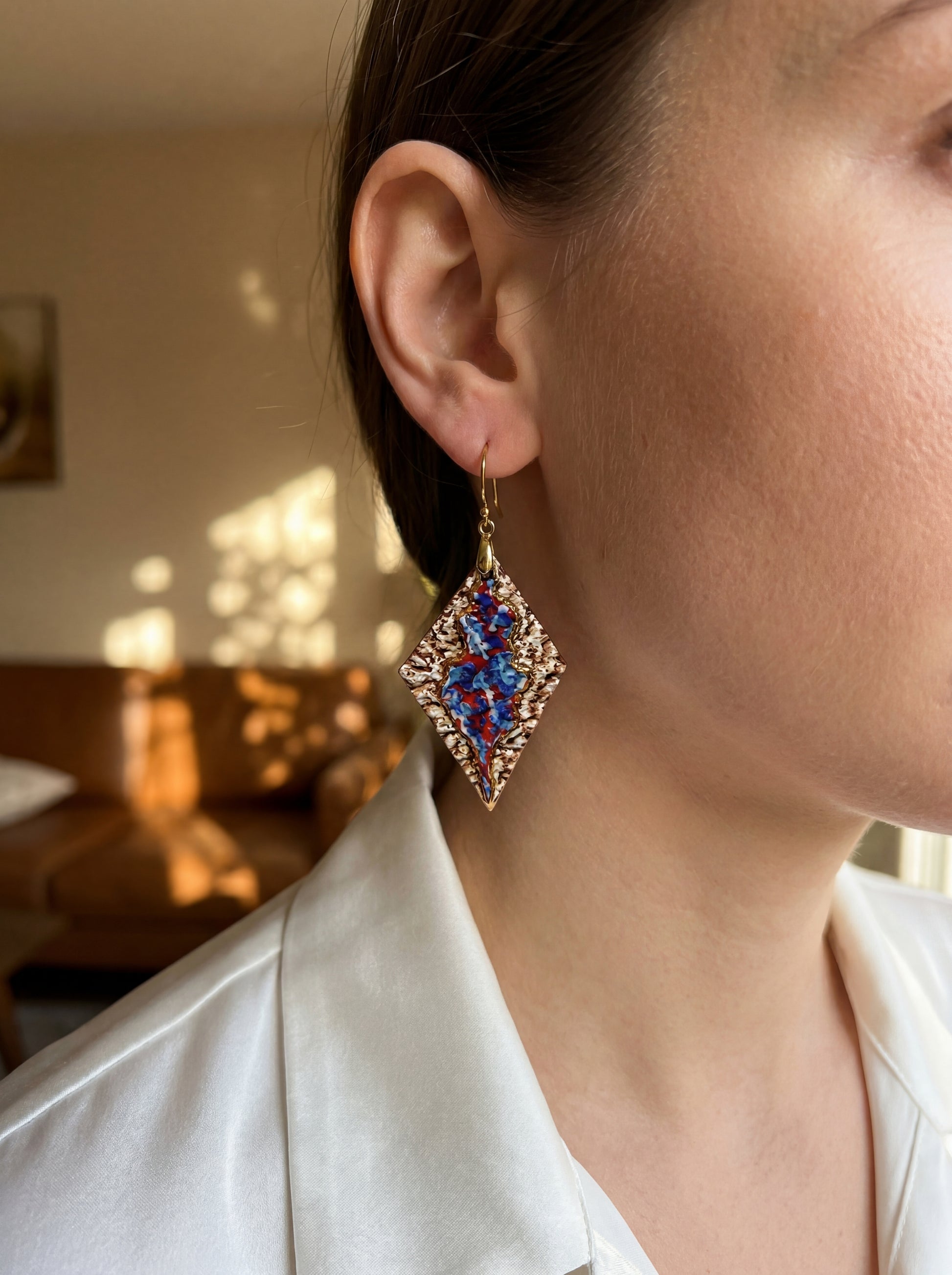 A model wearing HECROWN 005 handcrafted rhombus ceramic earrings in a warm, sunlit interior. The vibrant blue and red glaze contrasts with the textured bronze surface, showcasing the sculptural scale and elegant presence for daily wear.