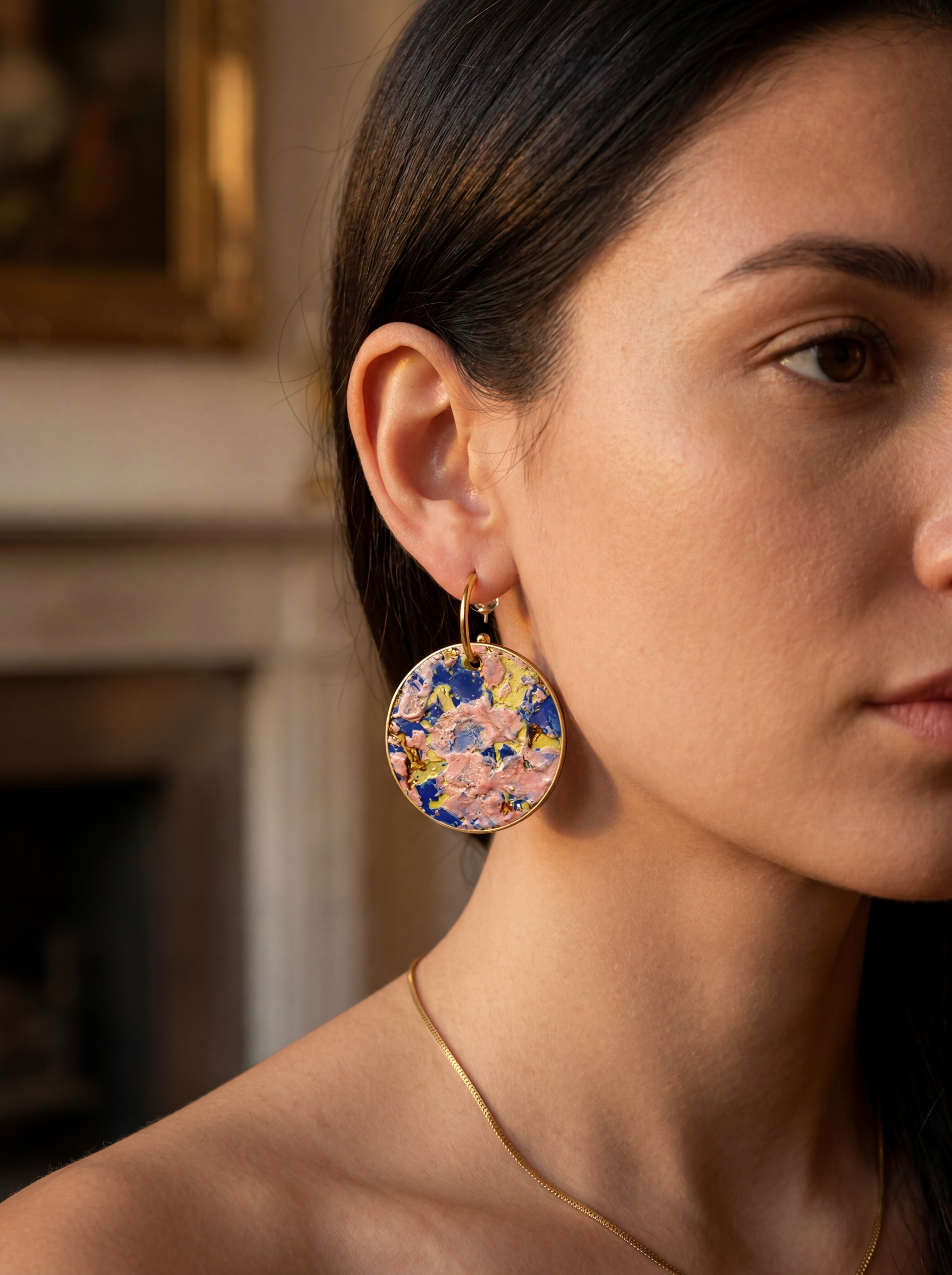 Side profile of a model wearing large round ceramic earrings with colorful impasto glaze textures in pink, blue, and yellow. HECROWN signature wearable art styled in a classic interior with an oil painting background, emphasizing an artistic and sophisticated lifestyle.