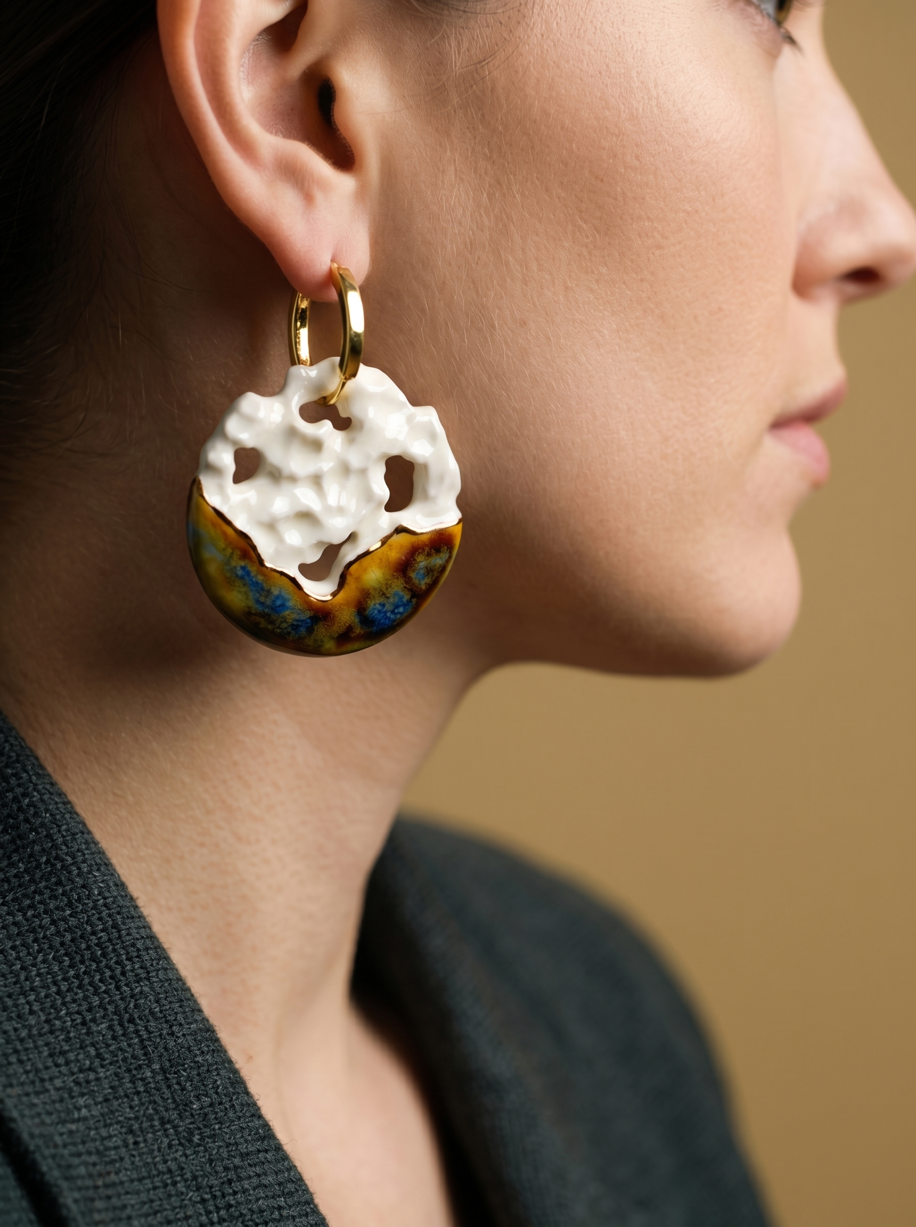 Side profile of a model wearing chunky handmade ceramic hoop earrings with a white crater-like texture and metallic brown glaze. HECROWN statement jewelry styled with a dark textured blazer, demonstrating the bold scale and quiet luxury aesthetic of wearable art