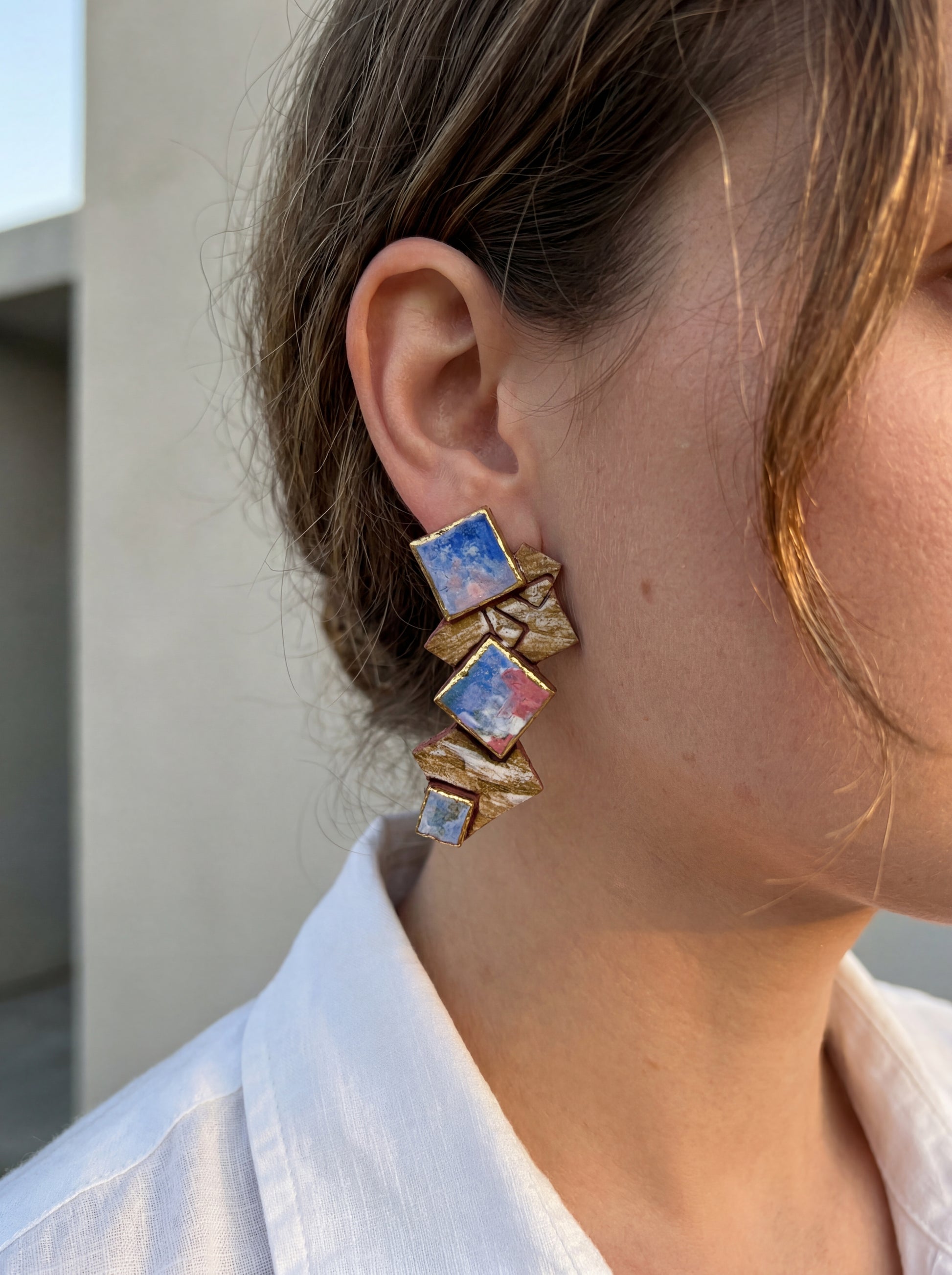 Side view of a model wearing HECROWN 003 geometric ceramic earrings with layered blue and pink abstract squares. Styled with a crisp white linen shirt in natural daylight to showcase the sculptural elegance and wearable art aesthetic from Jingdezhen