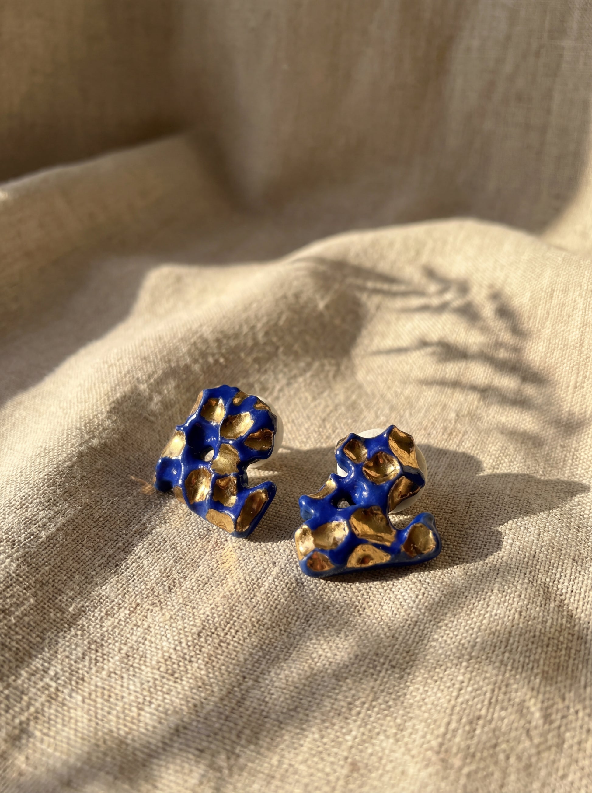 A pair of organic-shaped handmade ceramic stud earrings with deep cobalt blue glaze and shimmering gold accents. Displayed on a beige linen fabric under warm natural sunlight with botanical shadows, highlighting the sculptural texture and handcrafted soul of HECROWN's Jingdezhen artistry.